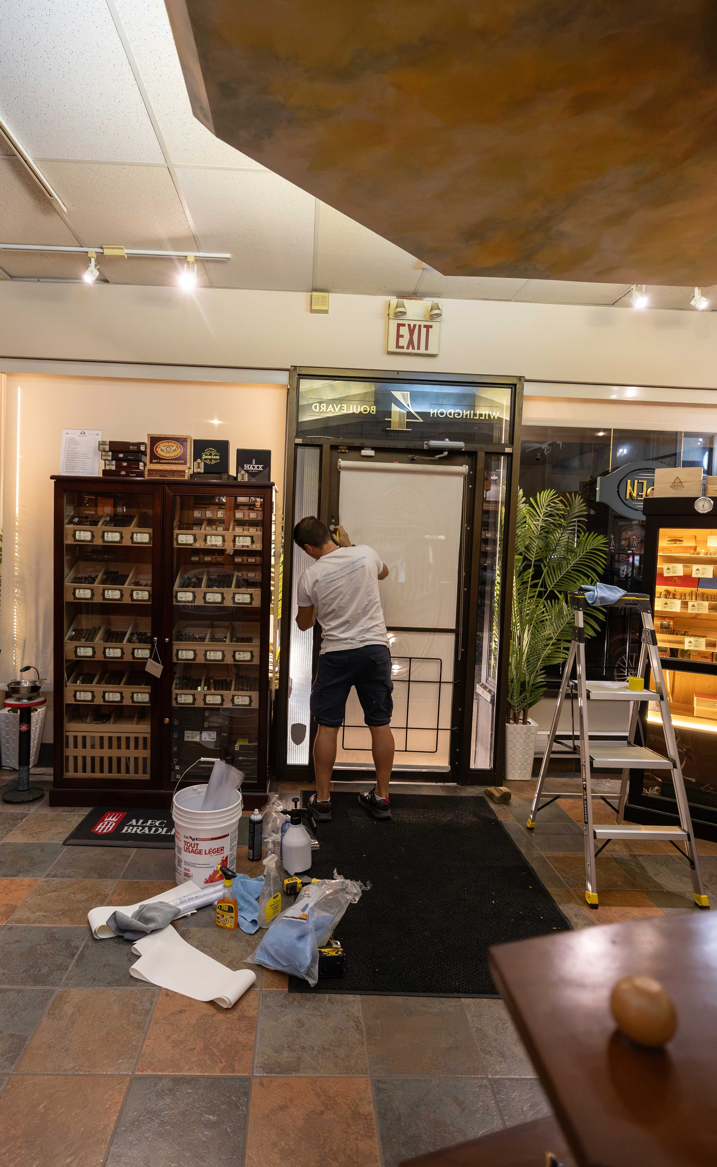 A worker installing or repairing a door in a commercial space. Tools and materials are scattered on the floor, including a bucket, spray bottles, and various hand tools. There is an exit sign above the door, and a glass display case with cigars or si