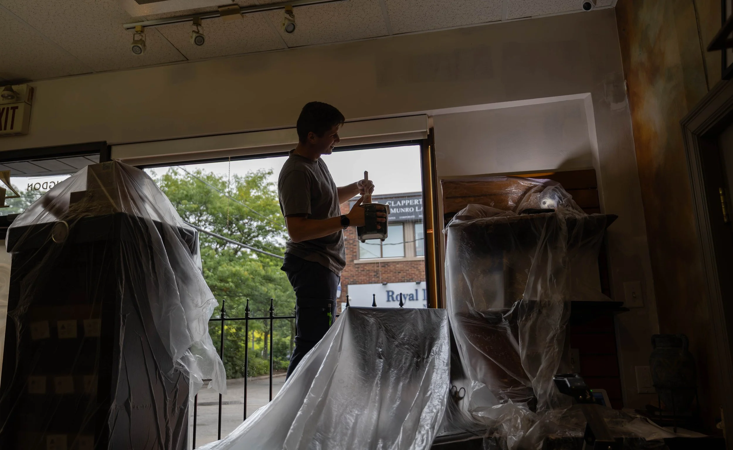 A man standing near a large open window, holding a drink and stirring with a straw inside a coffee shop or restaurant. The window reveals an urban street scene with trees and building signs outside.