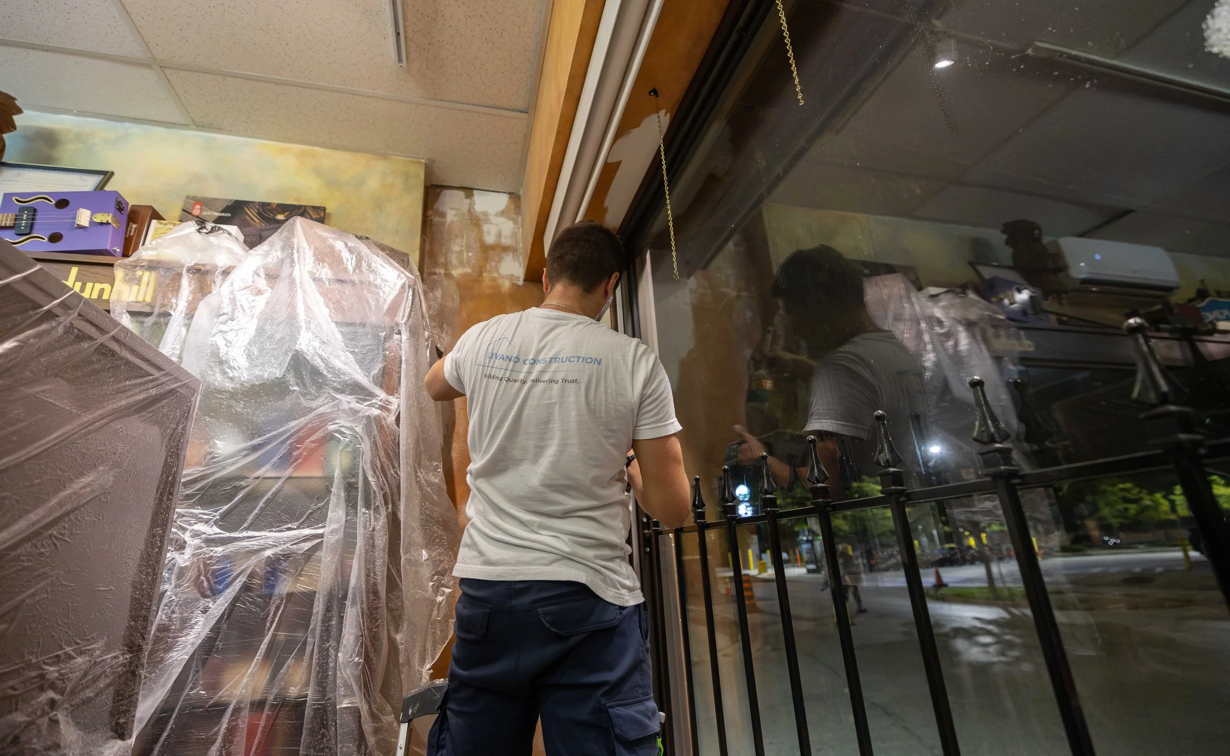 A person wearing a white T-shirt with company logo and name 'LIVAND CONSTRUCTION' on the back, standing inside near a large window, working on or inspecting the window area. The interior features a wall with various items like a purple guitar-shaped 