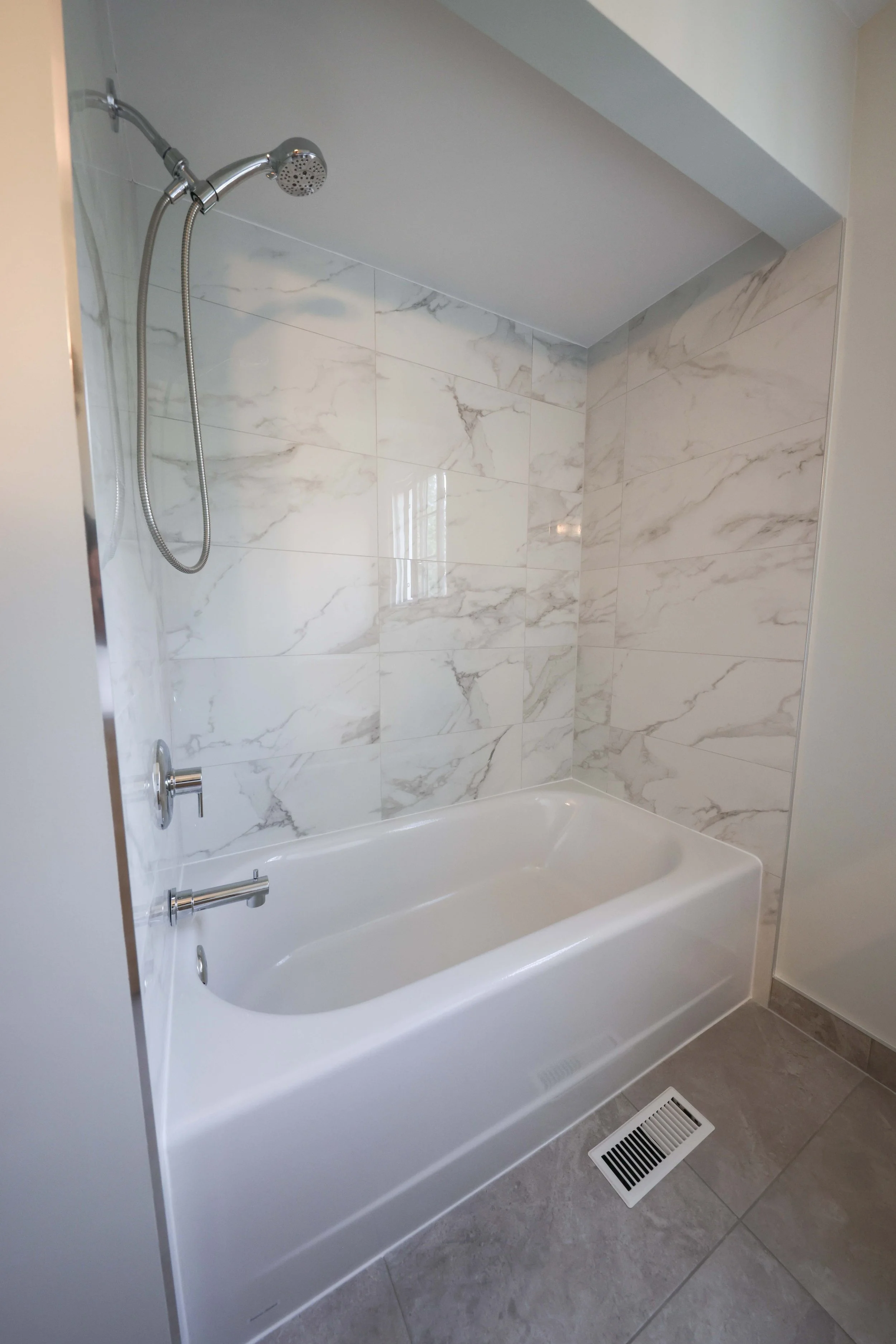 Bathroom with bathtub, marble tiles on walls, chrome showerhead on a flexible hose, drain and vent on floor.