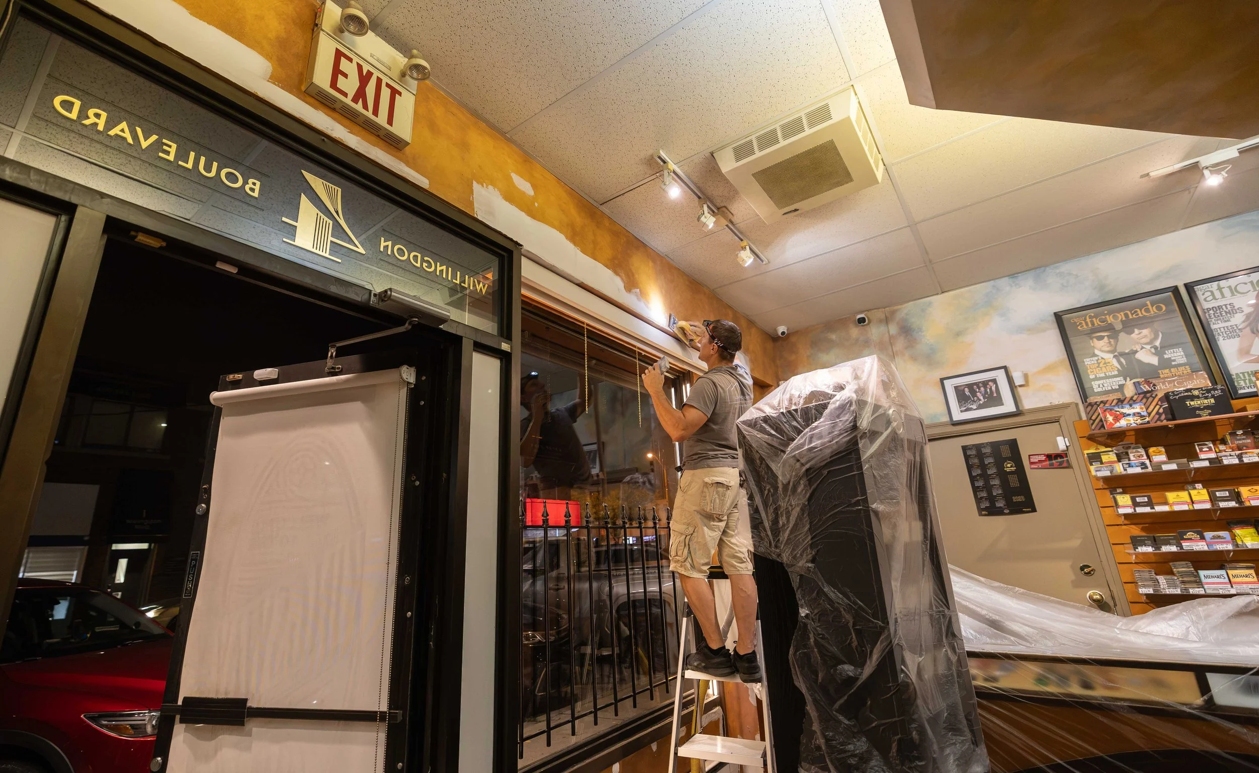 A person wearing a mask and casual clothing is cleaning a large window in an indoor space. The space has a ceiling with a ventilation unit and track lighting, and wall decorations including posters and a framed photograph. An exit sign is visible nea