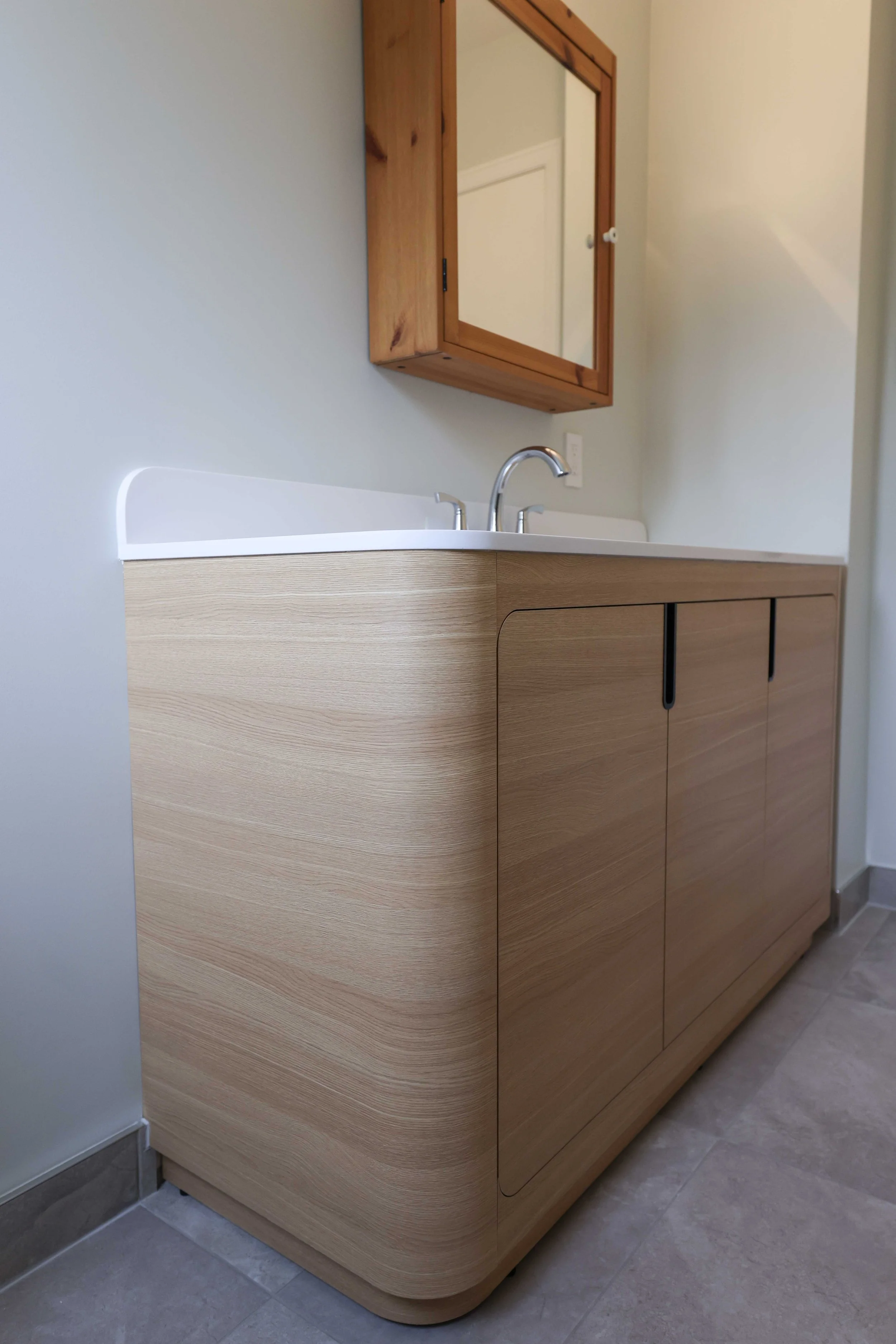 A bathroom vanity with a light wood finish, curved edges, and black handles, topped with a white countertop and a stainless steel faucet. A wooden framed mirror cabinet is mounted above the sink on a light-colored wall.