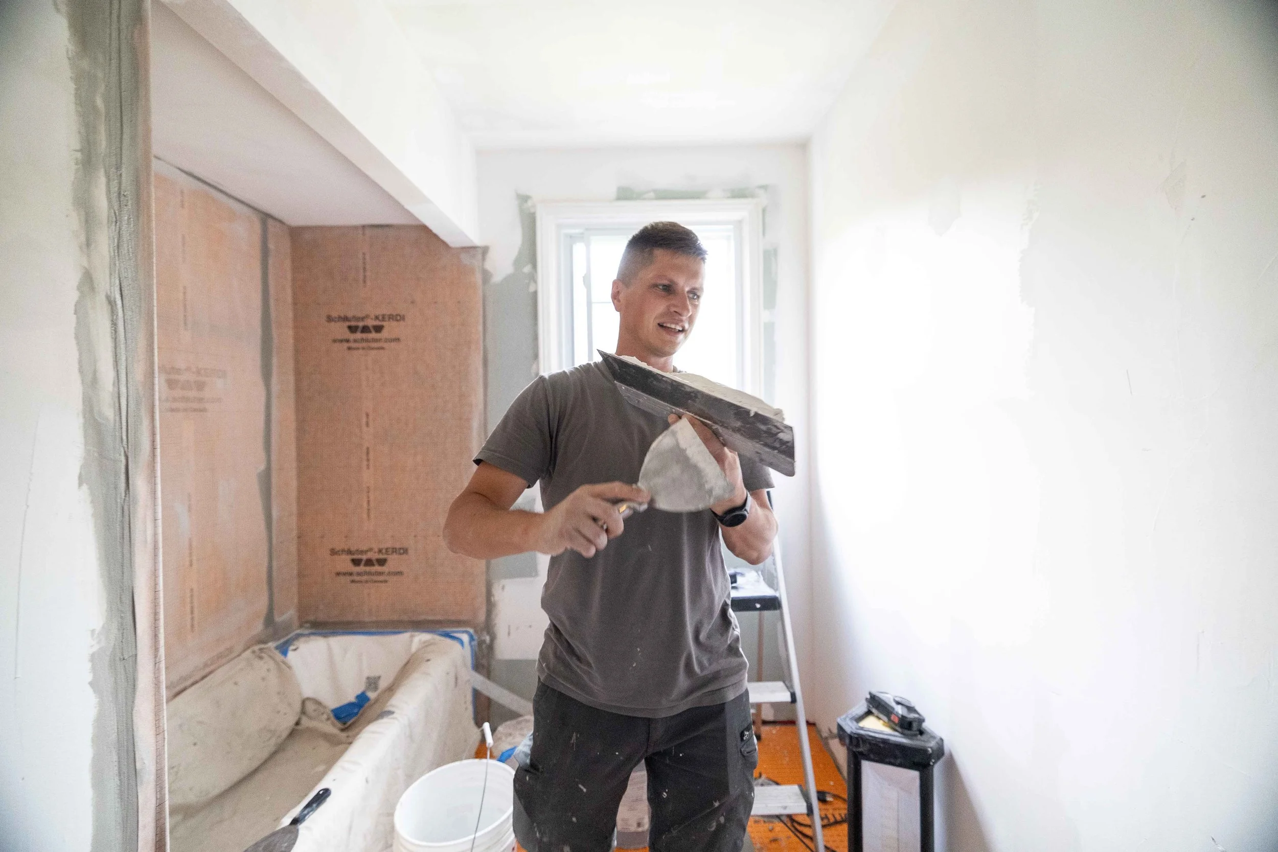 A man installing drywall in a room under renovation, holding a drywall knife and standing in front of a partially finished wall.