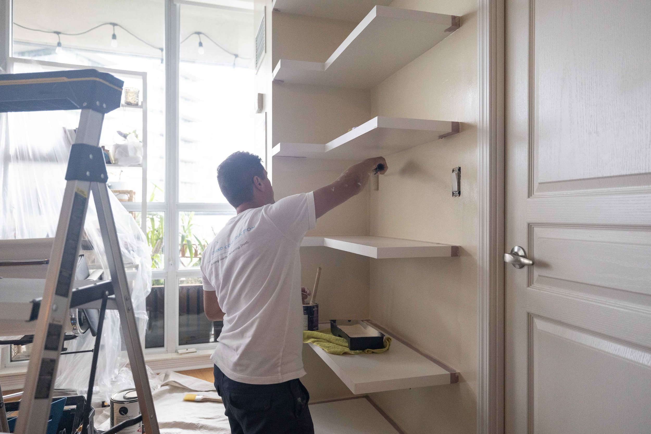 A man painting shelves inside a room, standing near a white wall with a closed door, with a window in the background and a ladder nearby.