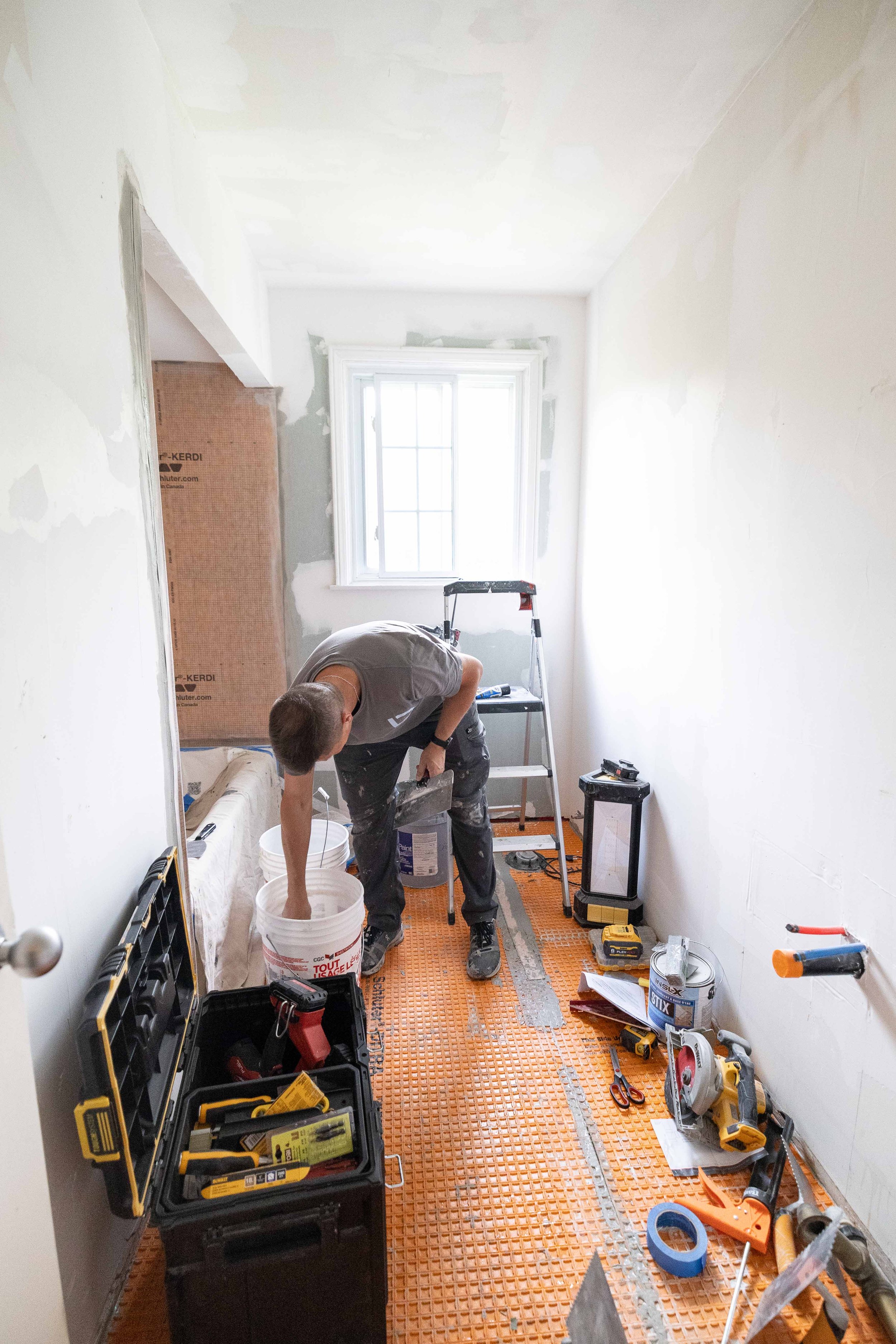 A person working on a home renovation in a room with white walls and a window. The room is scattered with various tools, construction materials, and an orange protective floor covering.