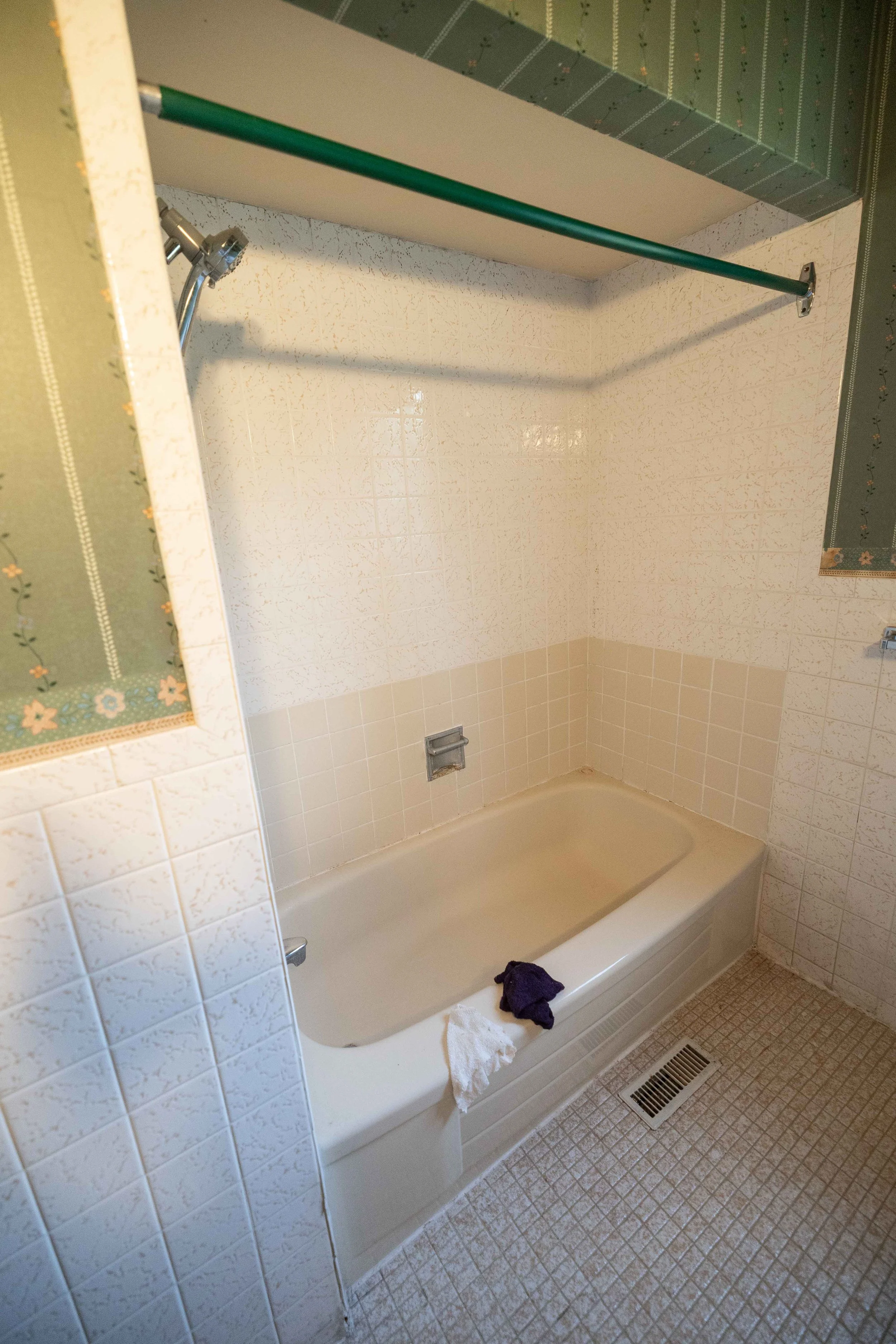 A bathroom with a bathtub, beige tile walls, and a small tiled floor area. On the bathtub edge are a black cloth and a white rag. The bathtub has an overhead shower with a flexible hose and a light-colored fixture, and a towel or cloth in the corner 