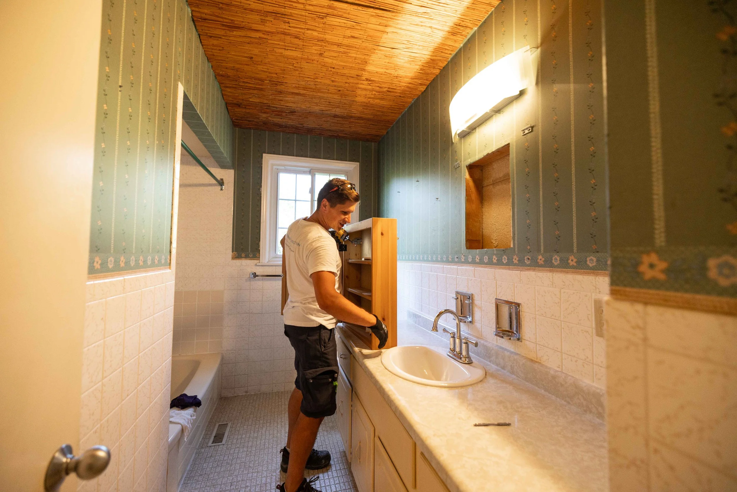 A man in a white t-shirt and shorts working on a cabinet in a bathroom with patterned wallpaper, a wooden ceiling, a window, and a sink.