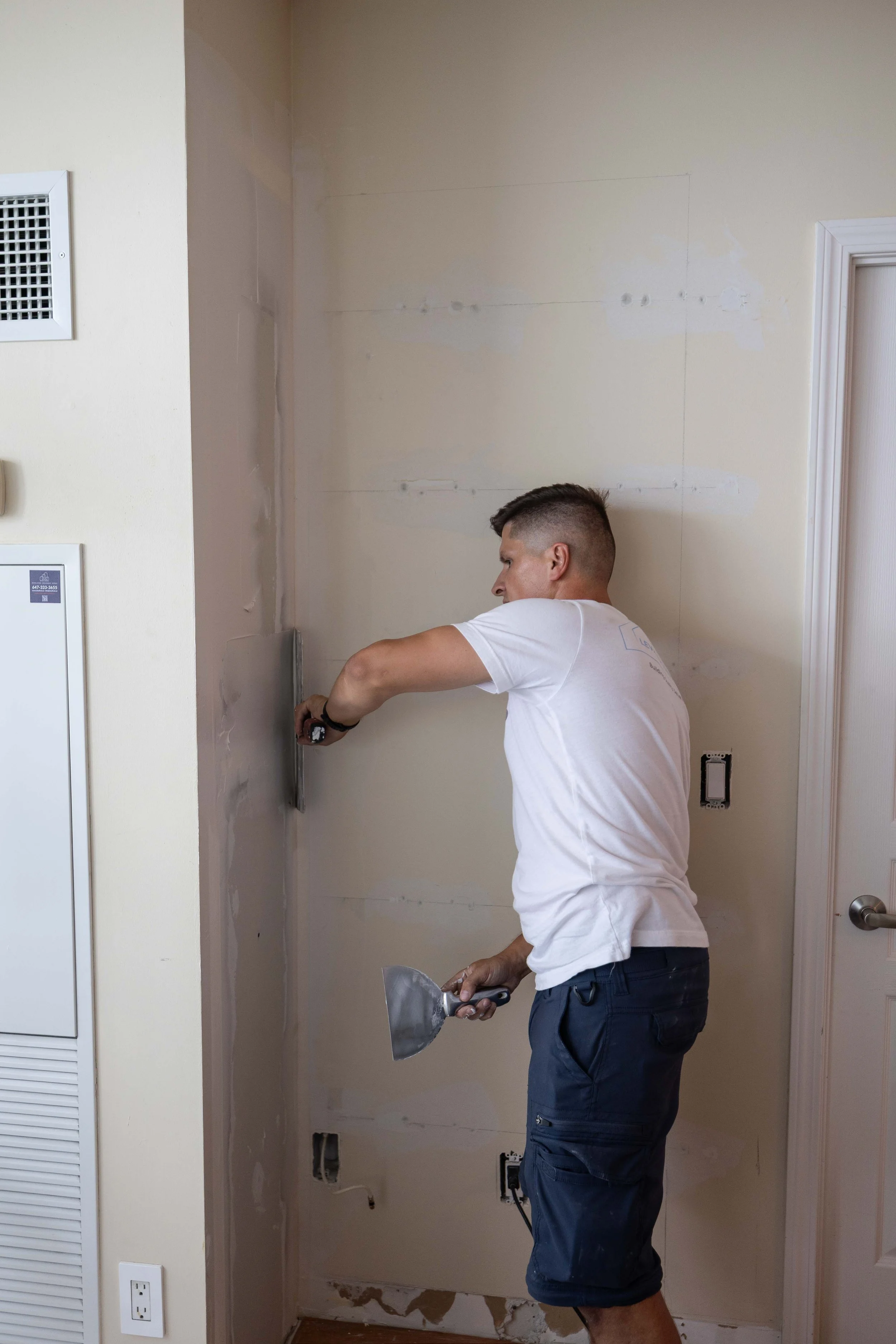 Man working on wall, using a trowel, in a room under renovation, with unfinished drywall and electrical outlets.