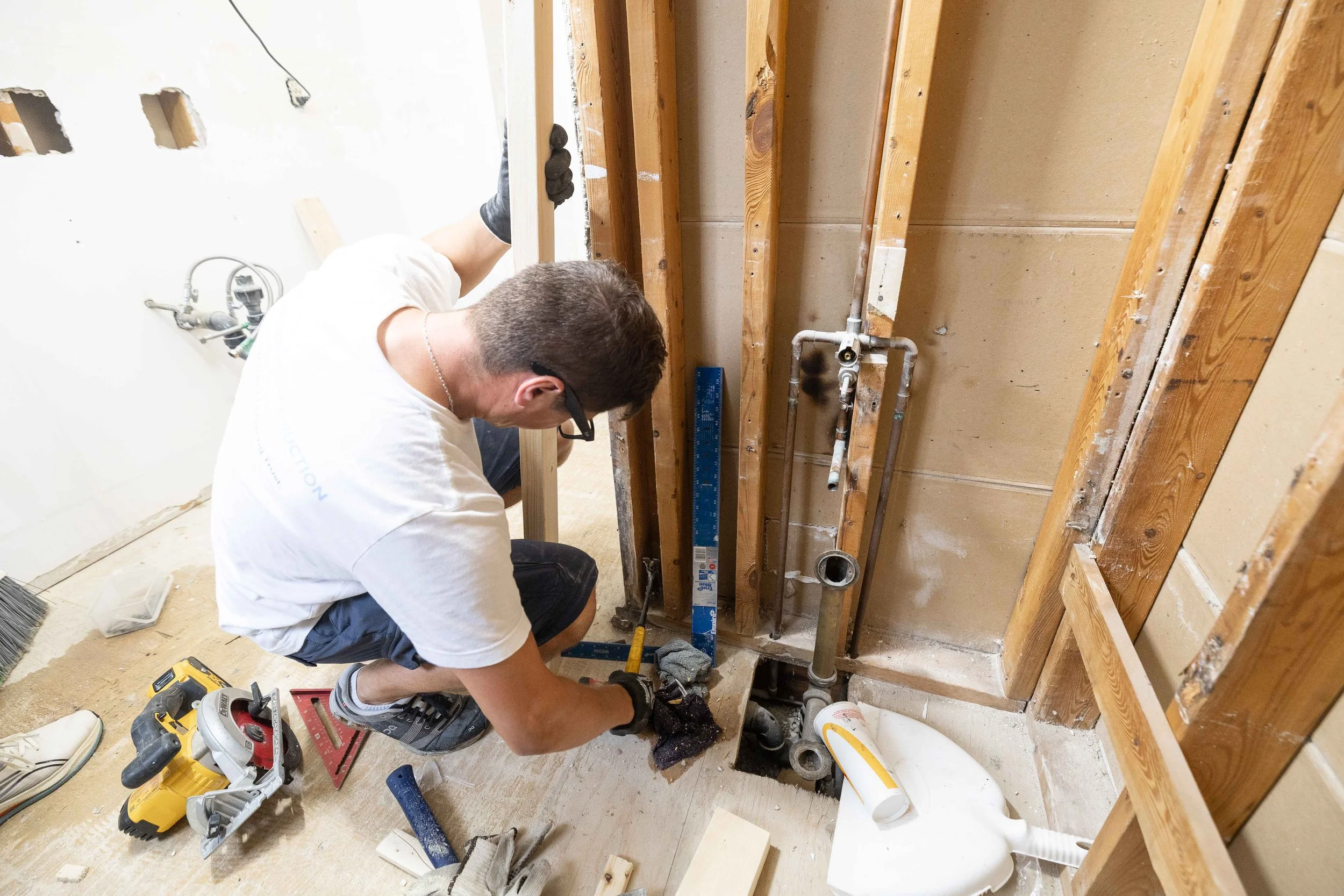 A person working on plumbing installation inside a partially open wall in a room under renovation, surrounded by tools, pipes, and construction materials.