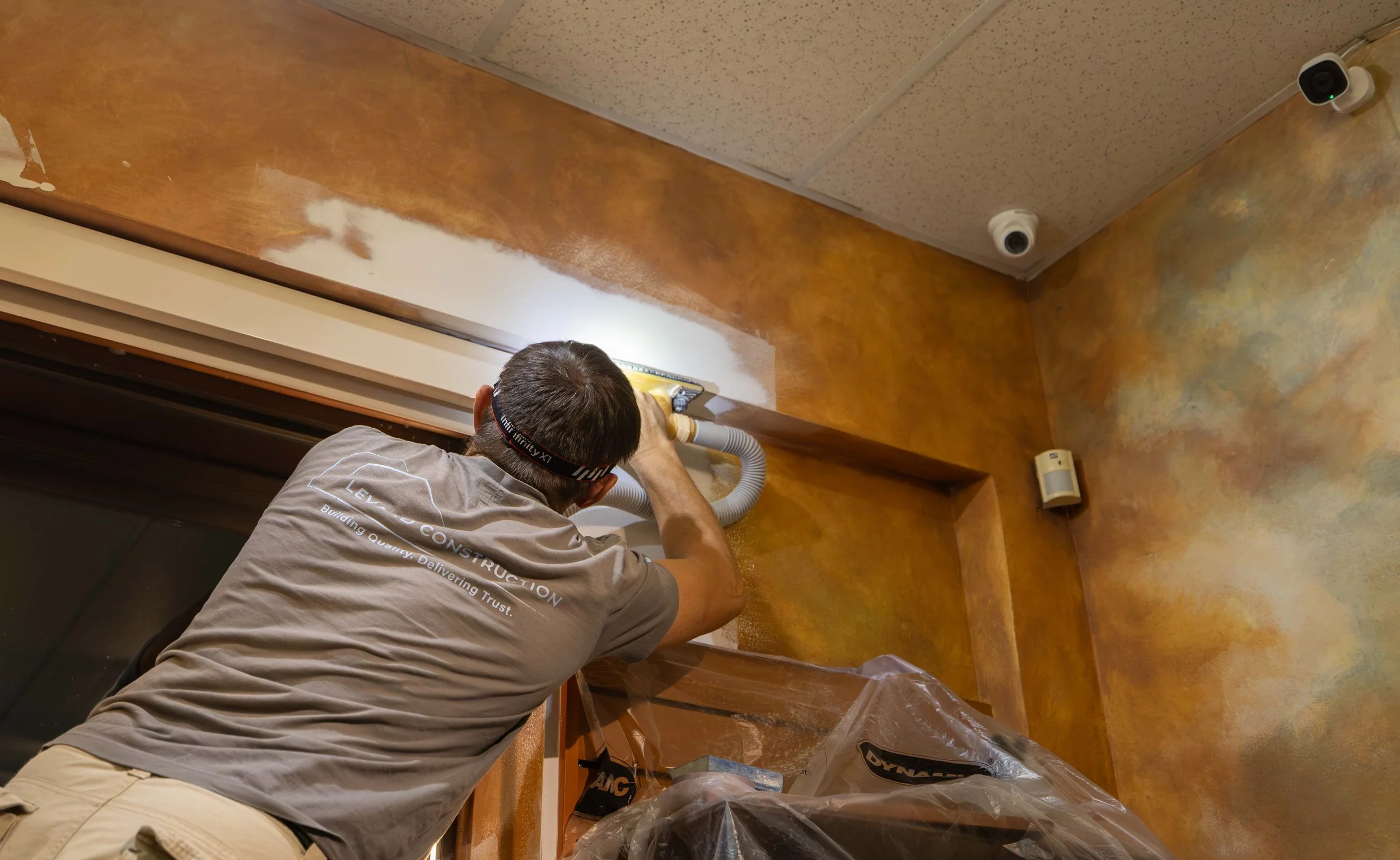 A person wearing a gray shirt and beige pants is vacuuming a corner of a room with brown and yellow textured walls and a wooden baseboard.