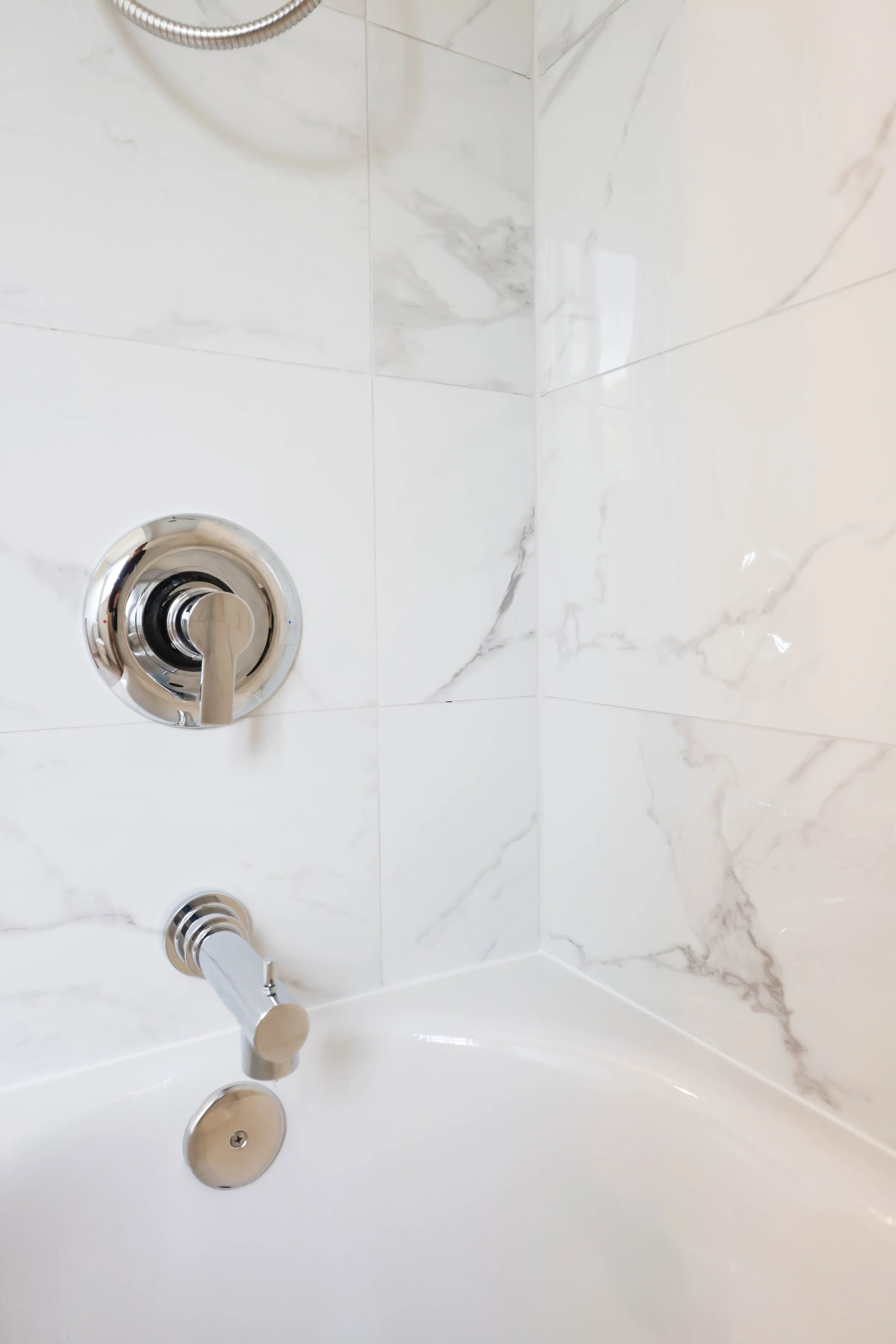 A close-up of a marble tiled bathroom shower with a silver shower handle, water flowing from the spout, and a drain on the white bathtub.