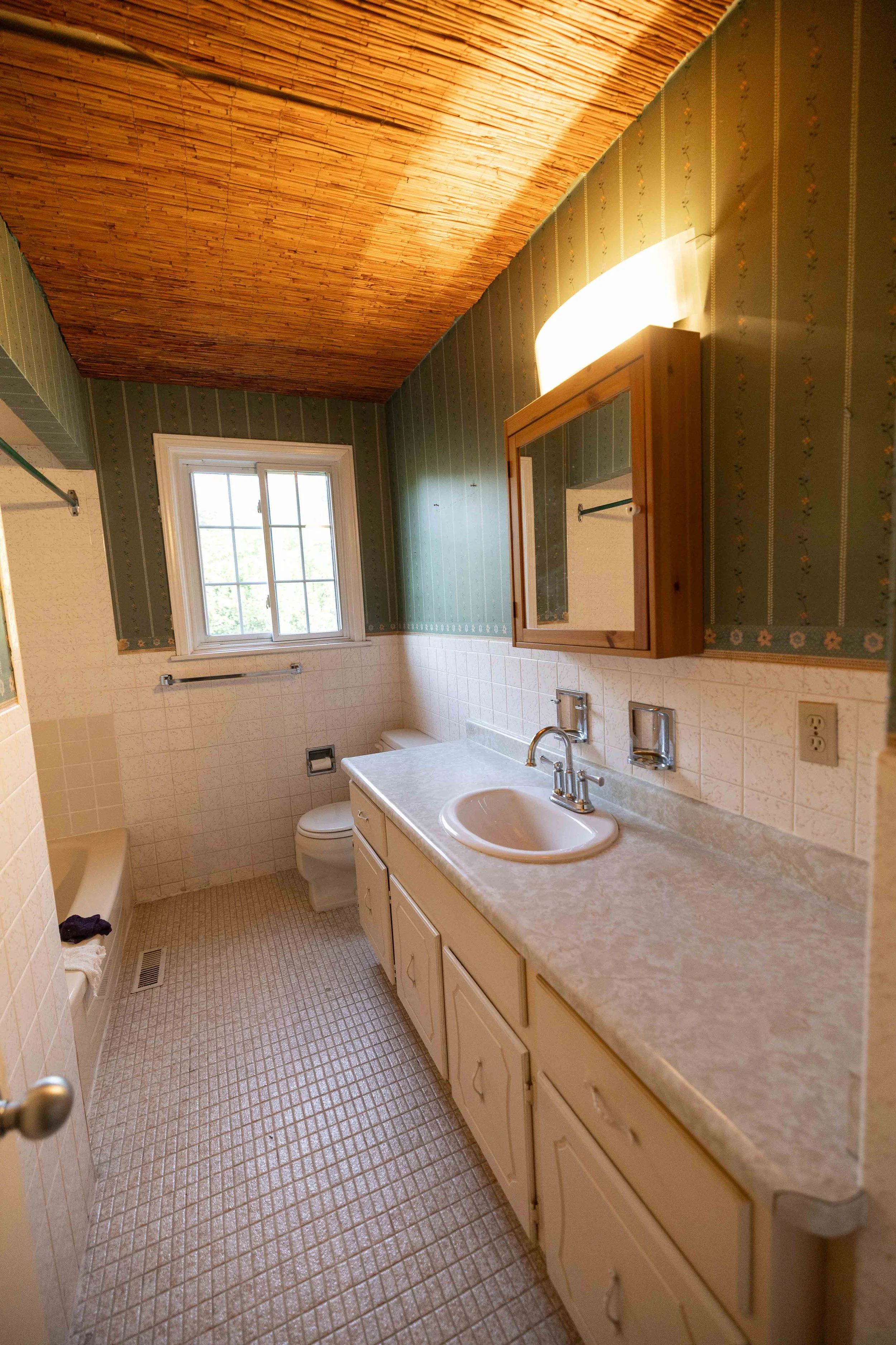 A bathroom with a beige vanity, marble countertop, and oval sink. There is a wooden medicine cabinet with a mirror above the sink. A window lets in natural light, and the ceiling has a wooden finish. The walls are decorated with patterned wallpaper a