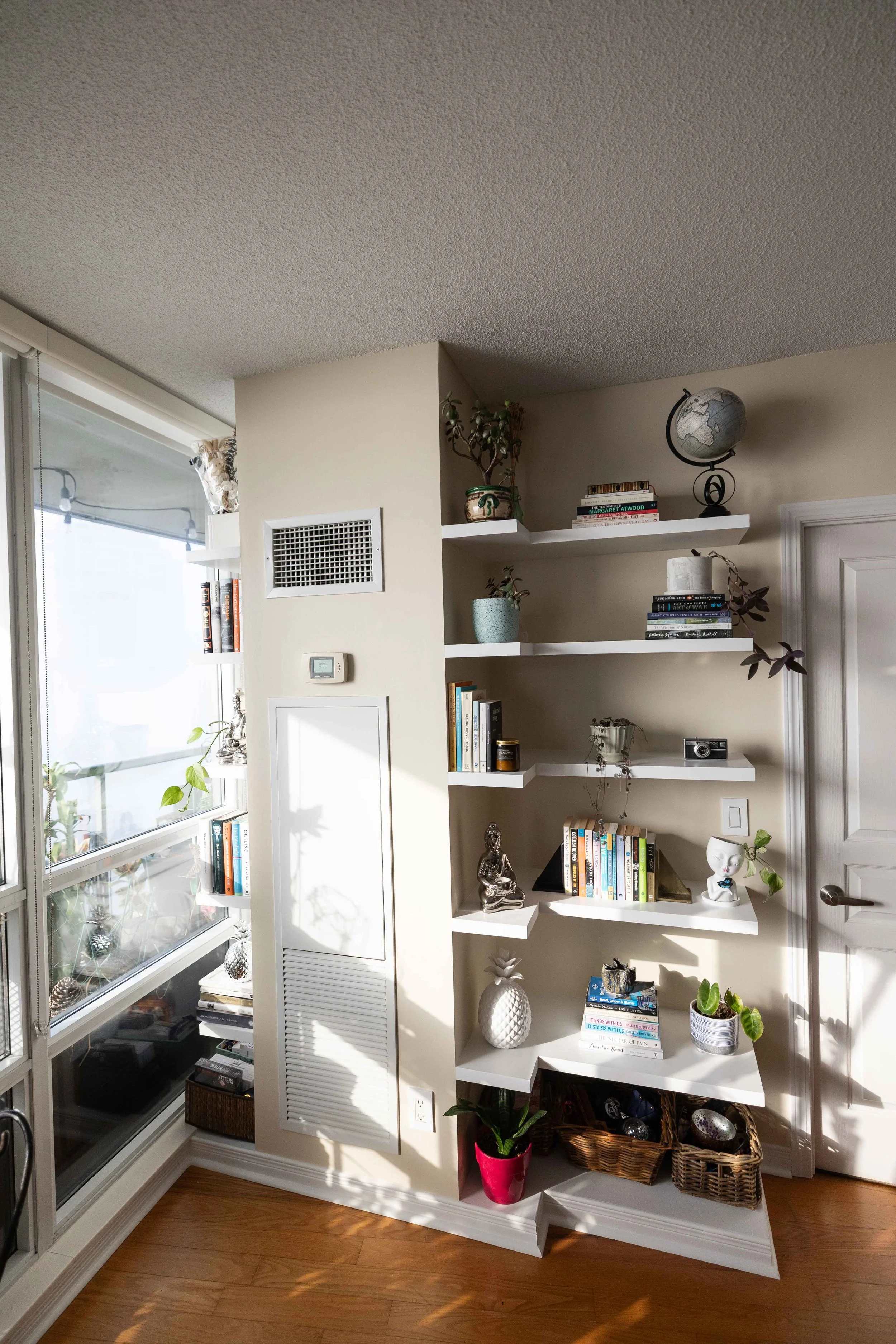 Interior view of a room with a white bookshelf, some decorative items, plants, and a window with blinds, sunlight casting shadows on a hardwood floor.