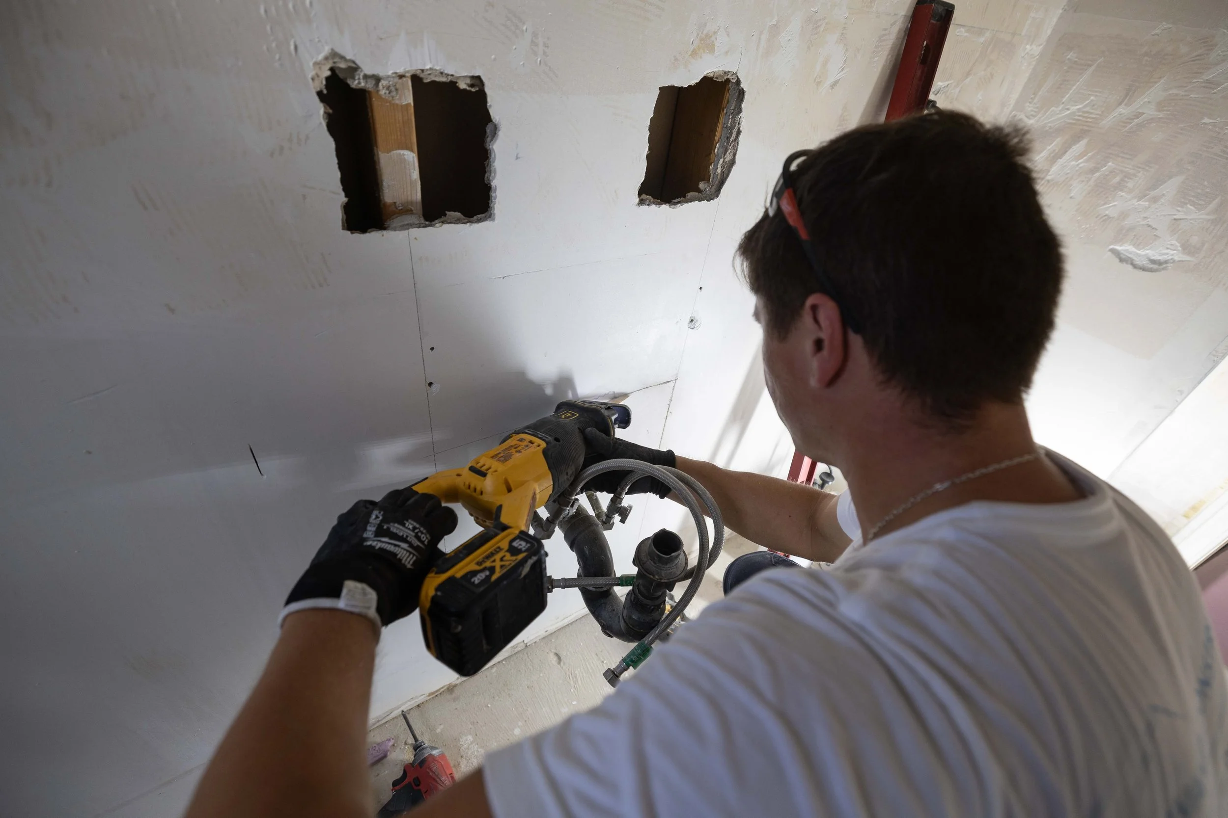 A person using a cordless drill to drill into a wall with two square holes. The person is wearing gloves and a white shirt, and is working on a construction or renovation project.