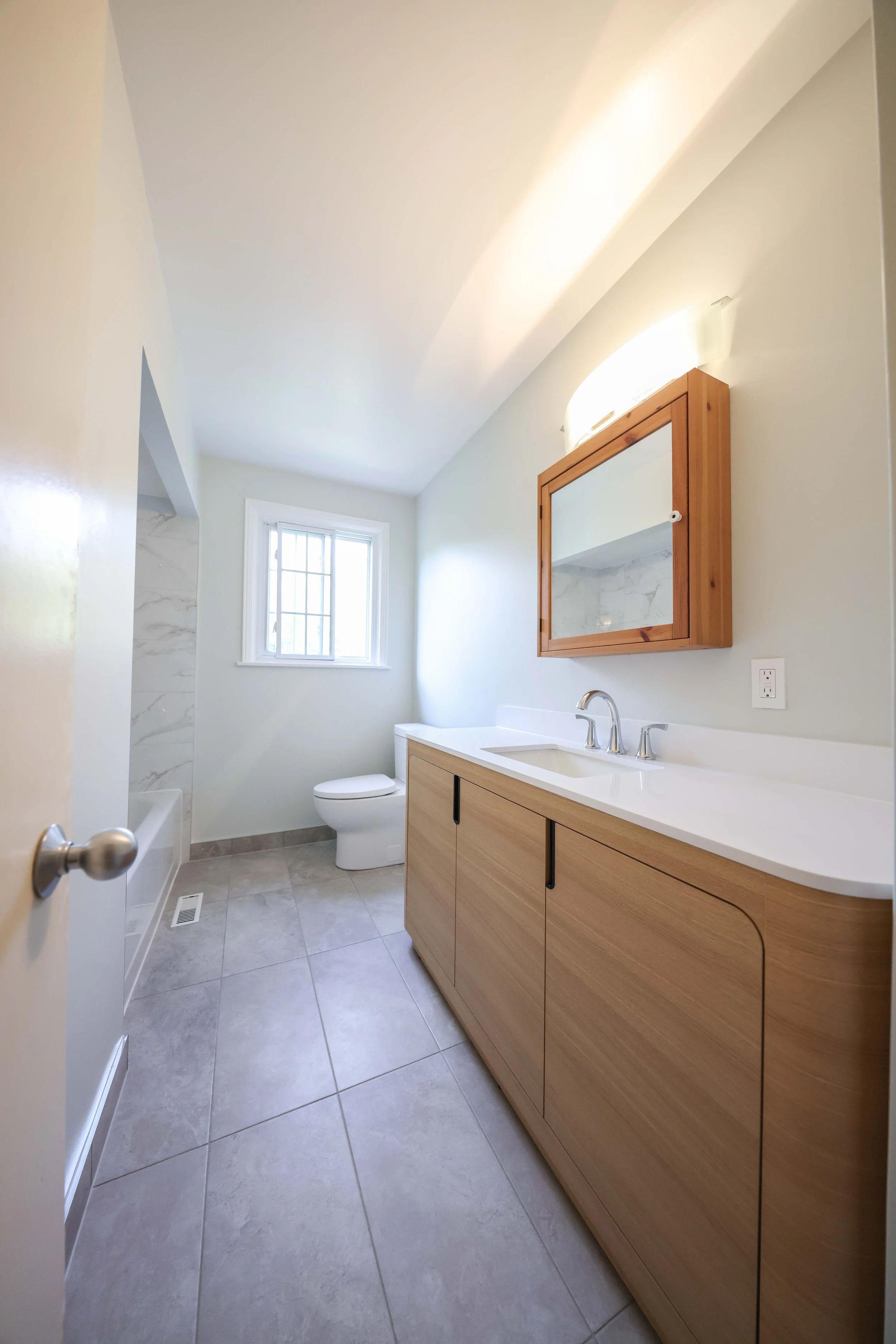 A modern bathroom with a large vanity featuring a white countertop and wooden cabinets, a rectangular mirror with a wooden frame, a single-handle faucet, a small window with grid panes, a toilet, and a tiled shower area with marble tiles. The floor i