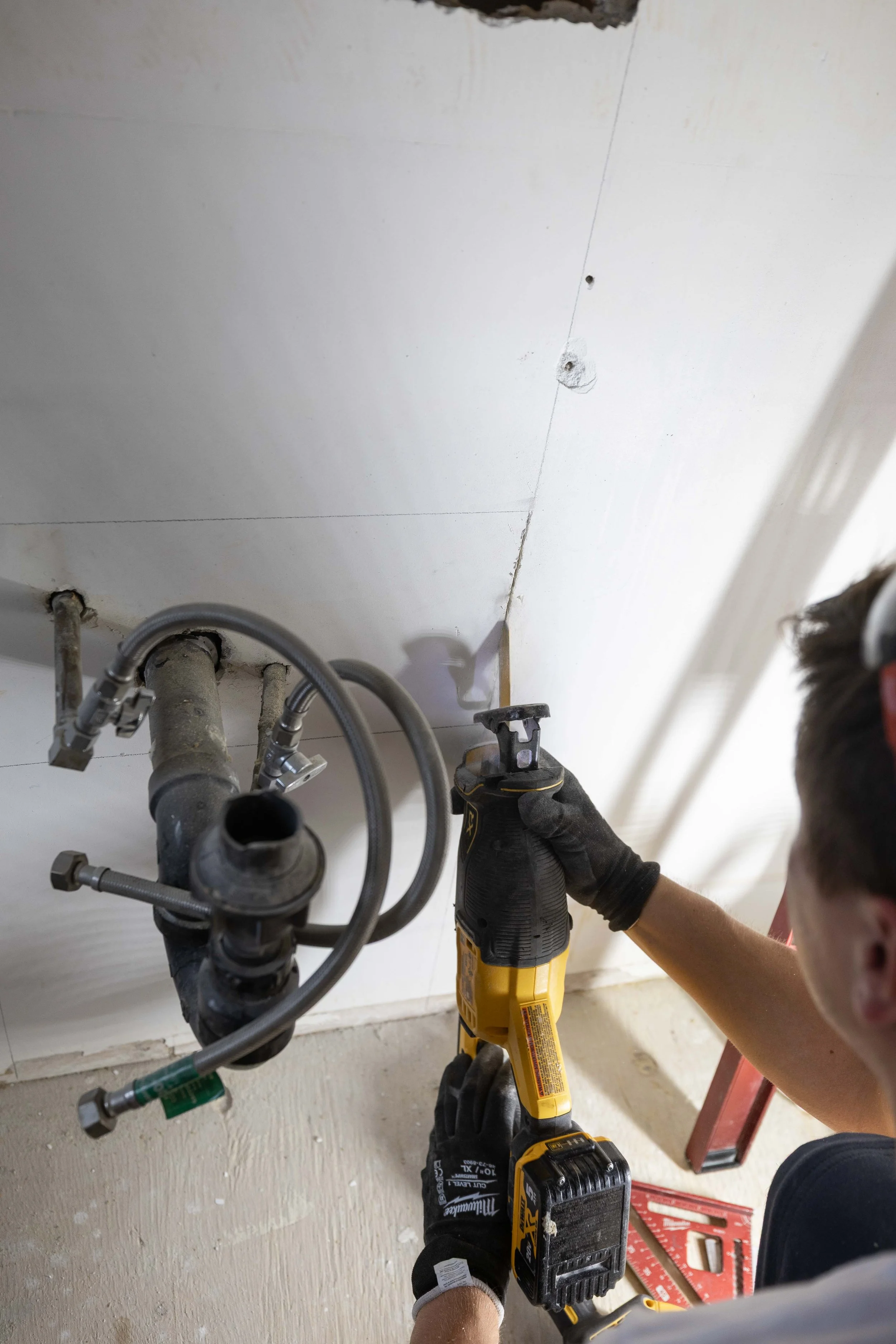 Person wearing black gloves using a yellow power drill to work on pipes underneath a white ceiling or wall.