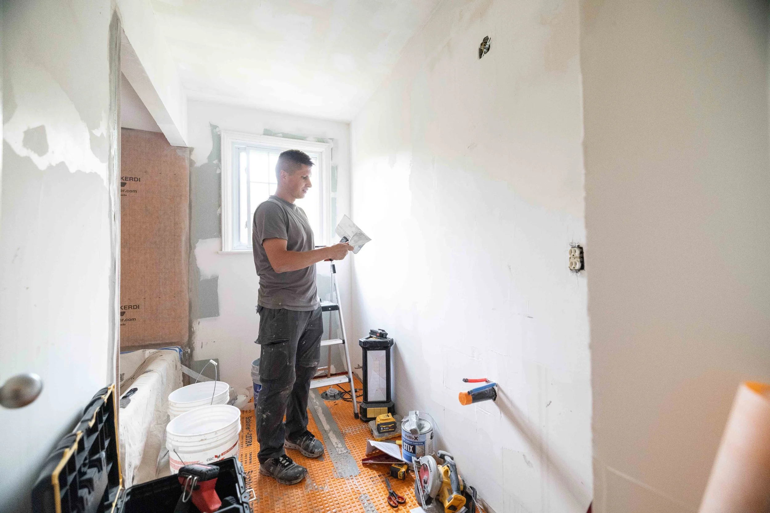 A man working on home renovation in a room with unfinished walls, tools, and construction materials around him.