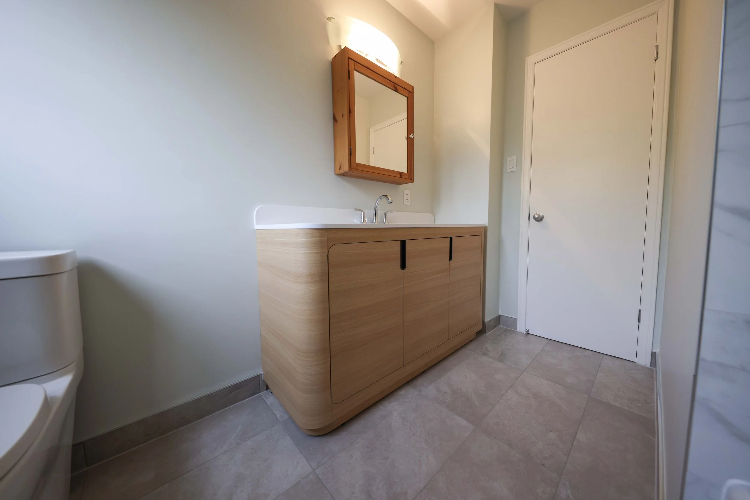 A modern bathroom with a wooden vanity, white countertop, and mirror, next to a white door and a partial view of a toilet on the left.