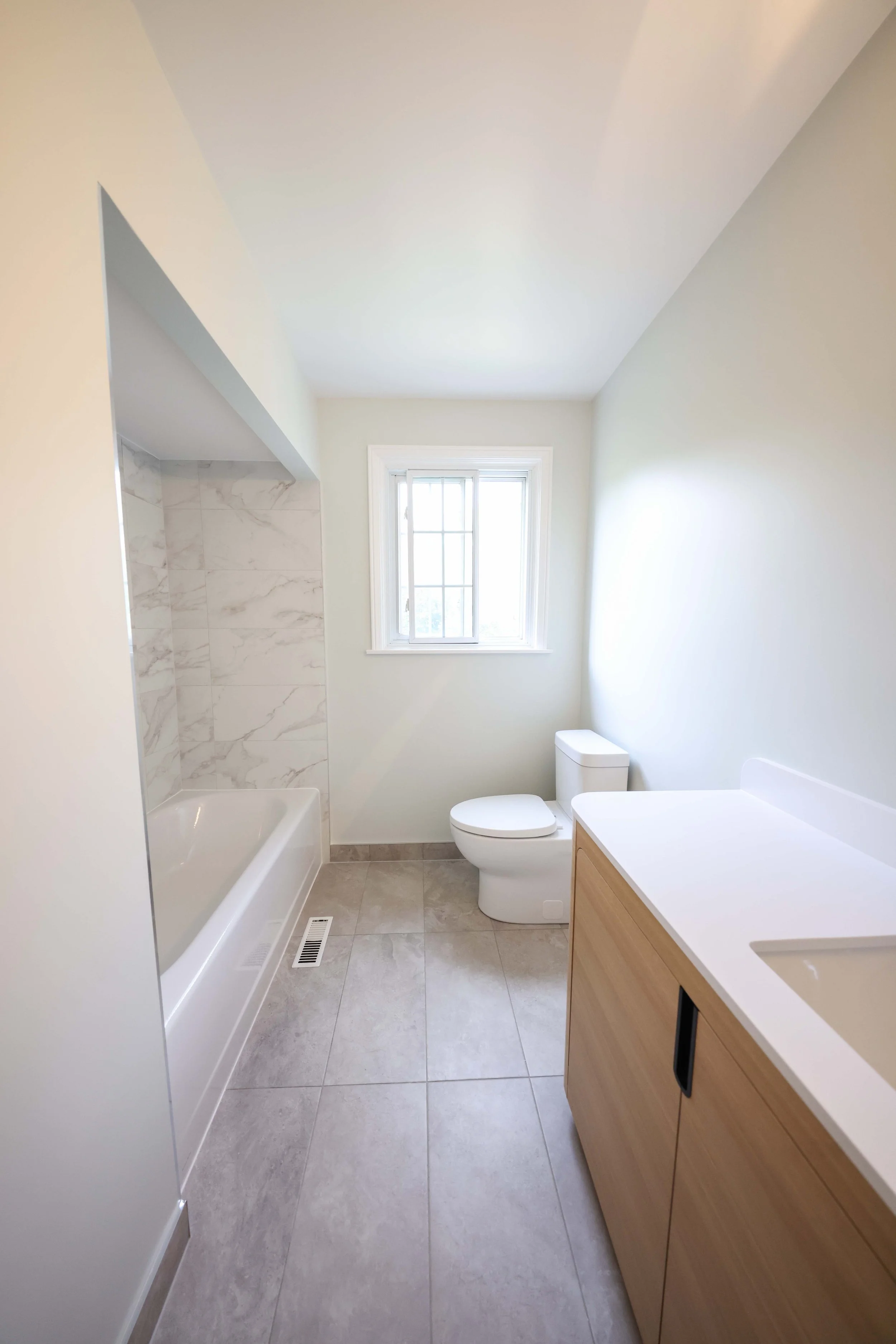 A modern bathroom with gray tiled floor, a toilet, a window, and a wooden vanity with a white countertop. There's a bathtub with a marble-tiled shower area and a vent on the floor.