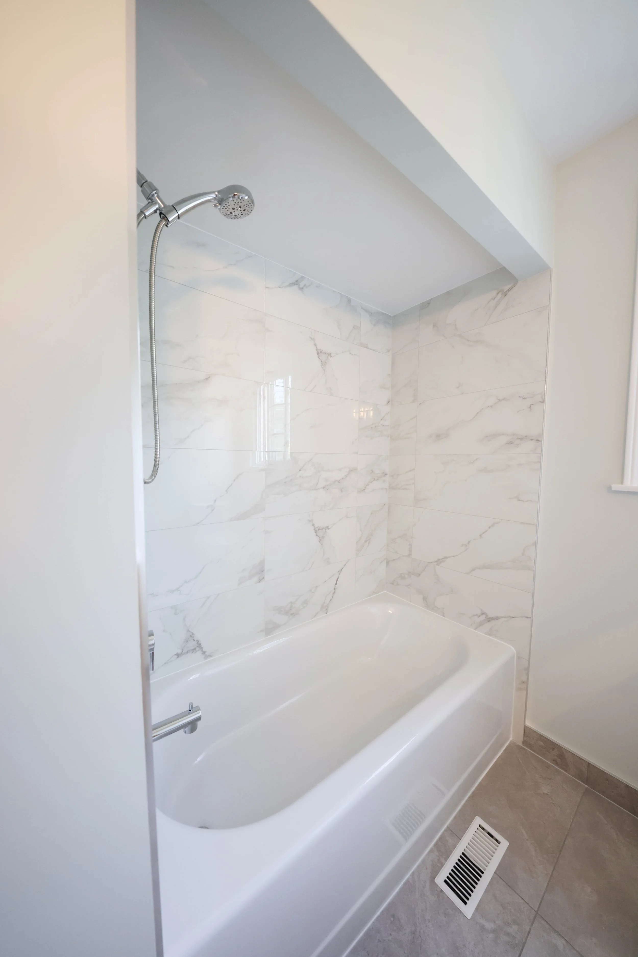 A bathroom shower with a chrome handheld showerhead, white marble tiles on the wall, a white bathtub, and a floor vent in the gray tiled bathroom floor.