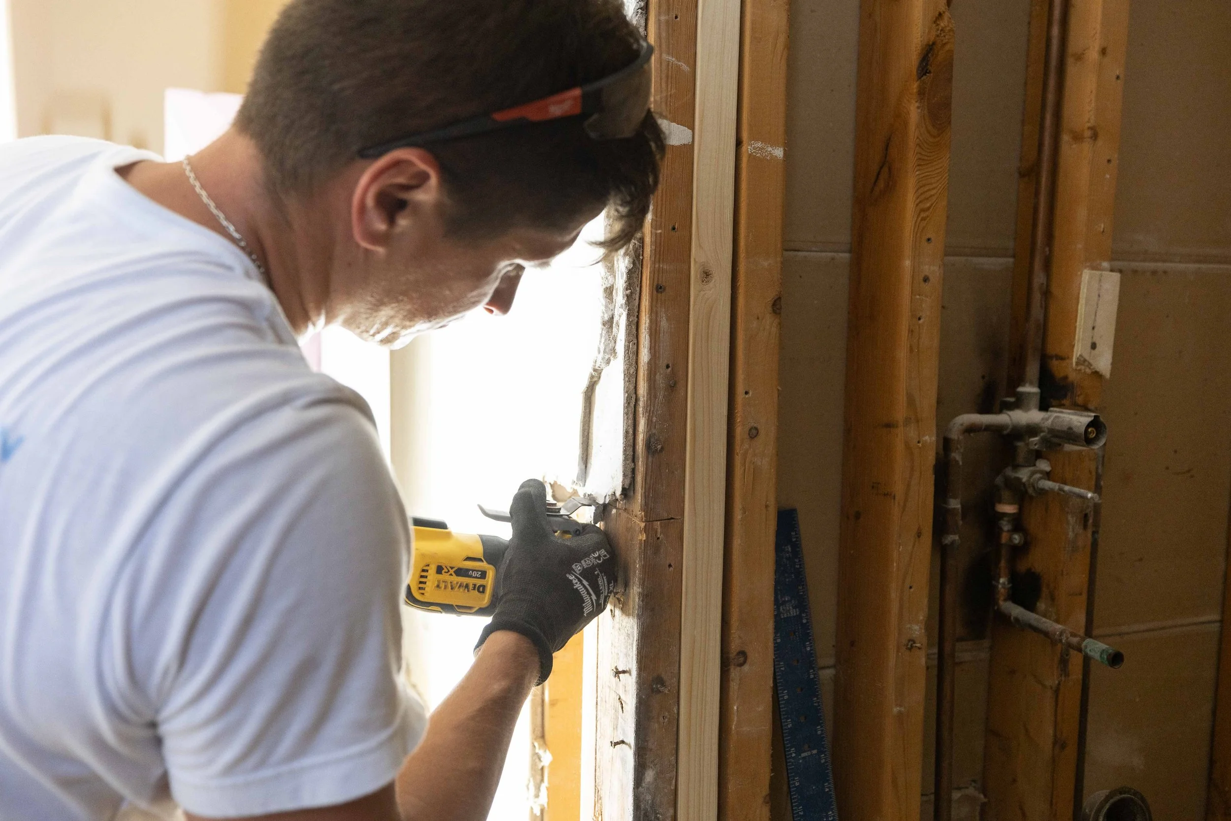 Man working on a construction project using a power drill, with exposed wooden framing and plumbing pipes visible.