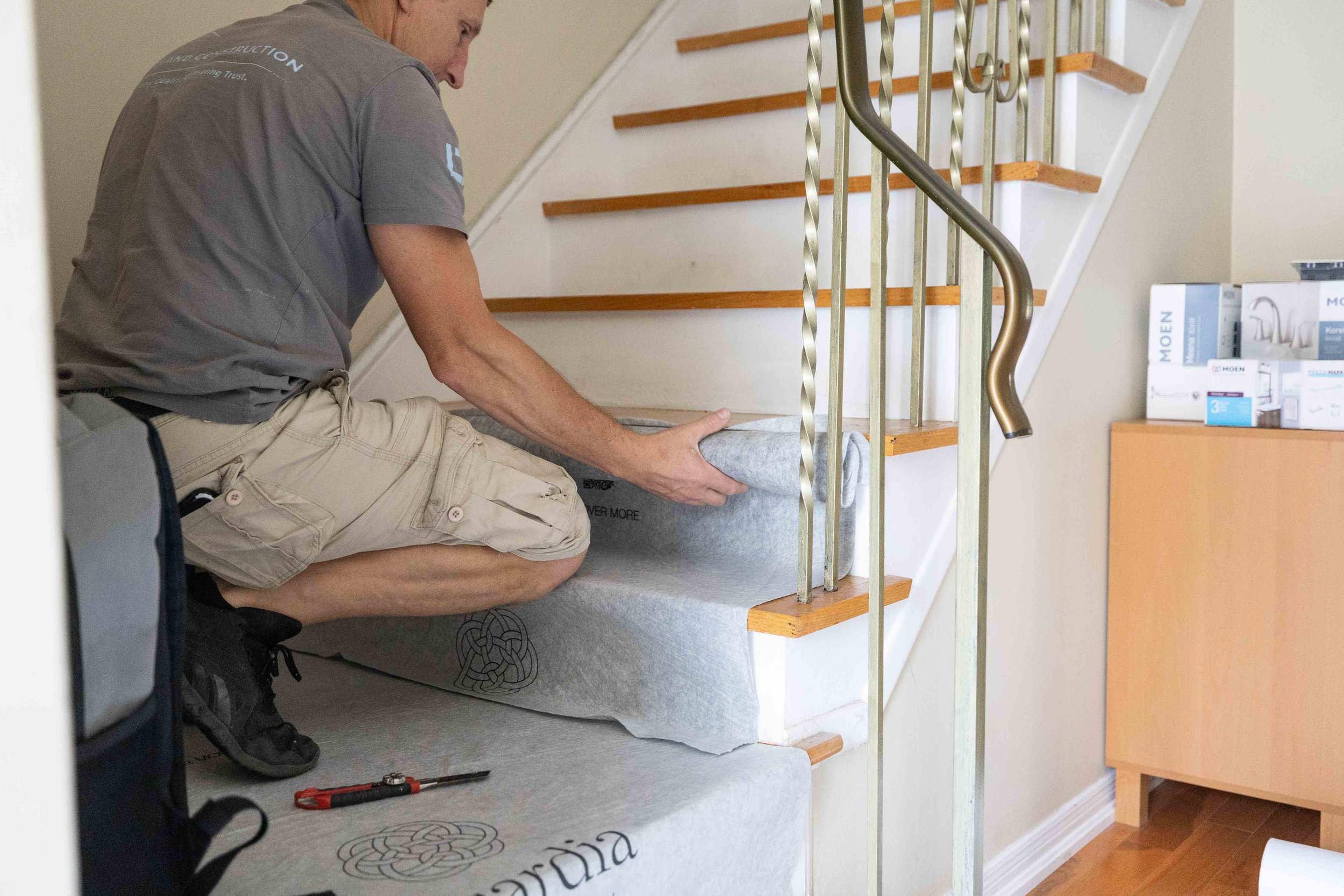 A man kneeling on a staircase carpet, installing or repairing it. He is holding a section of the carpet, and there are tools and boxed faucet products on a nearby table.