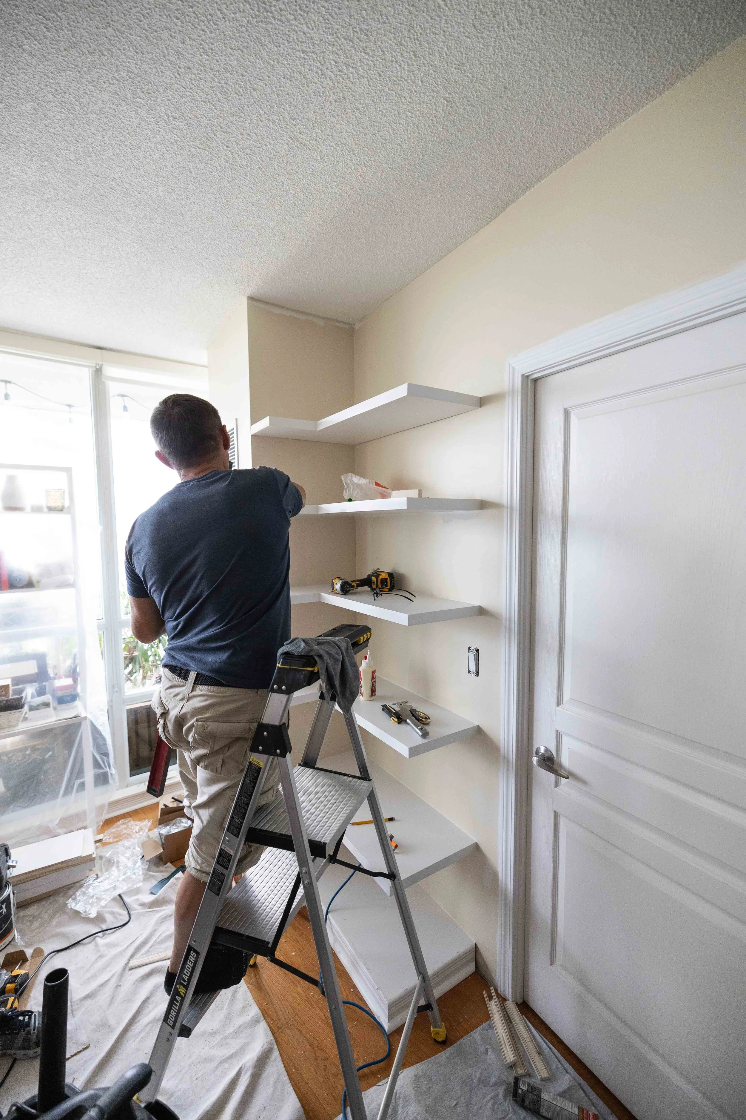 Person installing or assembling white floating shelves in a room, standing on a step ladder with tools on the shelves and floor, in front of a white door and a window with blinds.