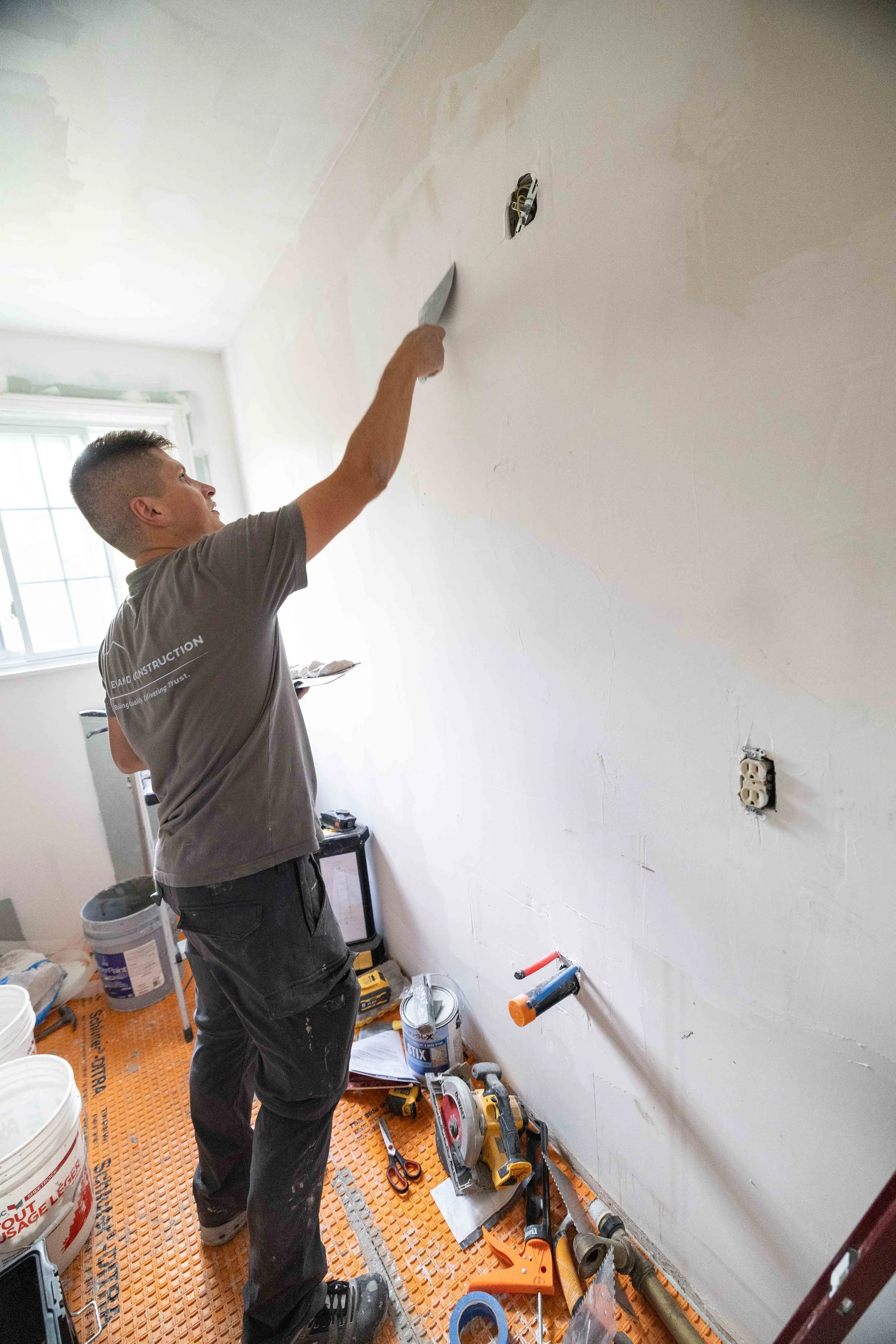 A person working on electrical wiring in a room with a partially finished wall. There are various tools, power drill, scissors, and construction materials on the floor.