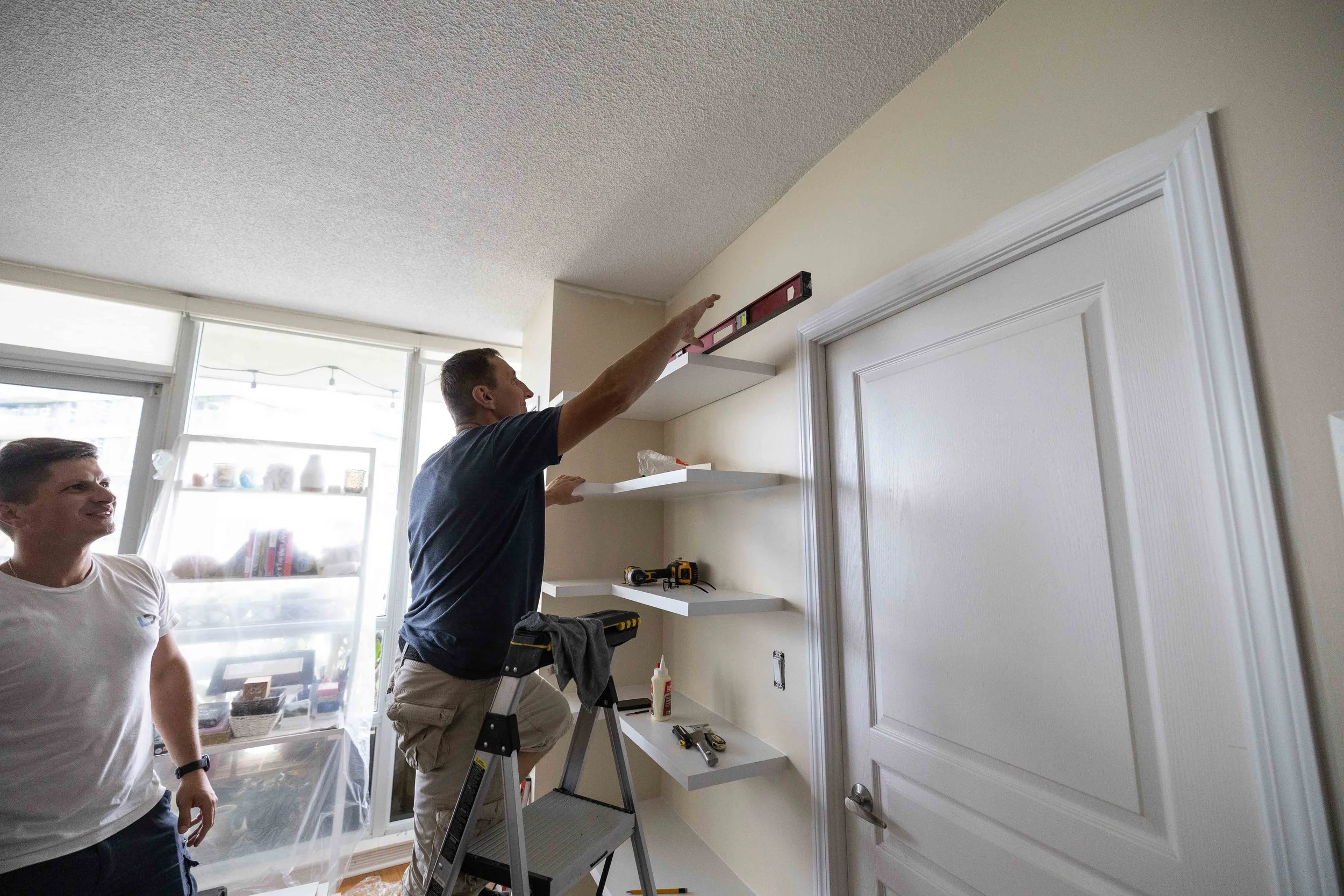 Two men installing white shelves in a room with a door and large windows. One man is standing on a ladder, pointing at the wall, while the other watches.
