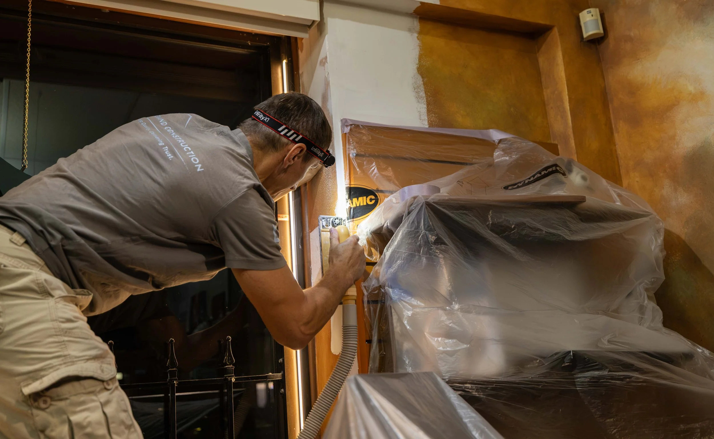 A person is working on a construction or renovation project, focused on a wall or surface with masking tape and protective plastic covering nearby.