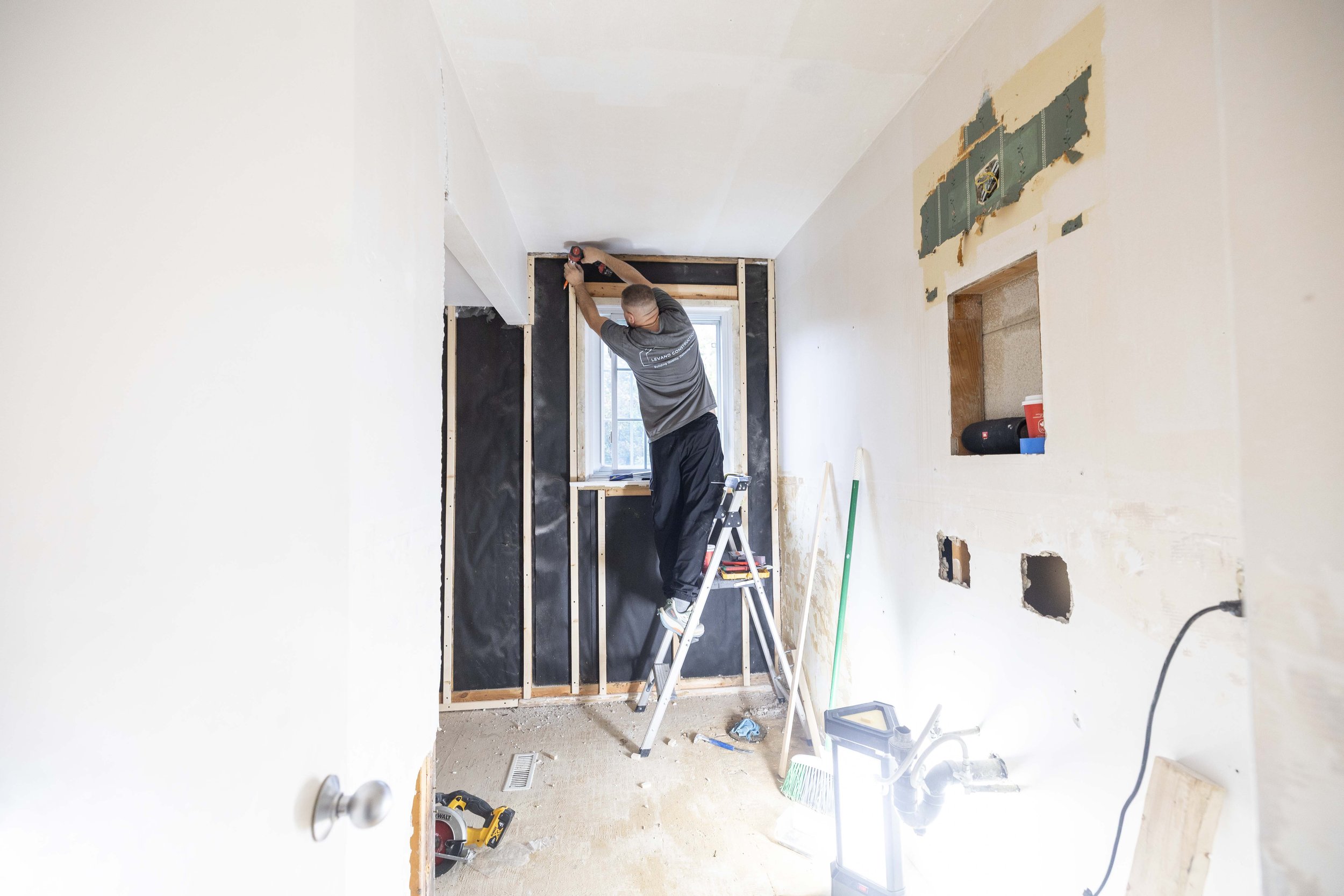 A person standing on a step ladder working on drywall in a room under construction, with tools and materials around.