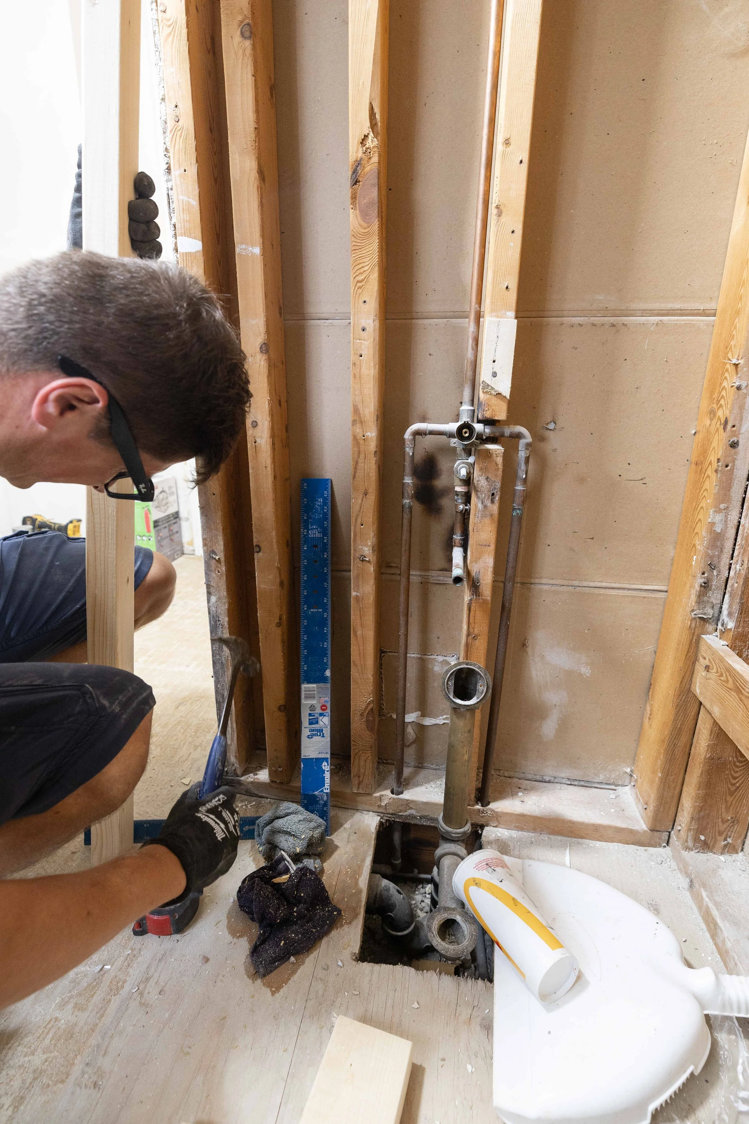 A person working on plumbing installation or repair inside a partially open wall with exposed wooden studs and pipes. Tools such as a level, a drill, and rags are visible on the floor.