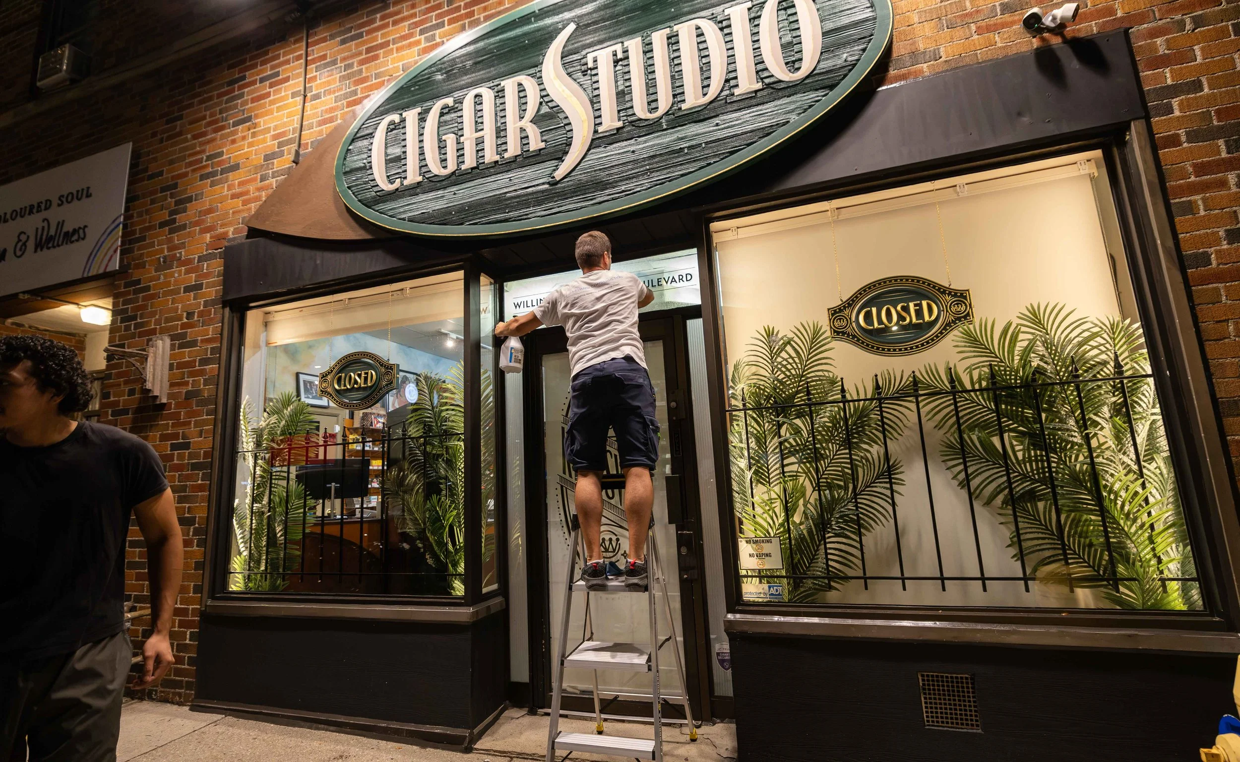Person standing on a ladder cleaning a large sign that reads 'CIGAR STUDIO' on a brick building. The building has a window display with plants and a sign indicating the business is closed.