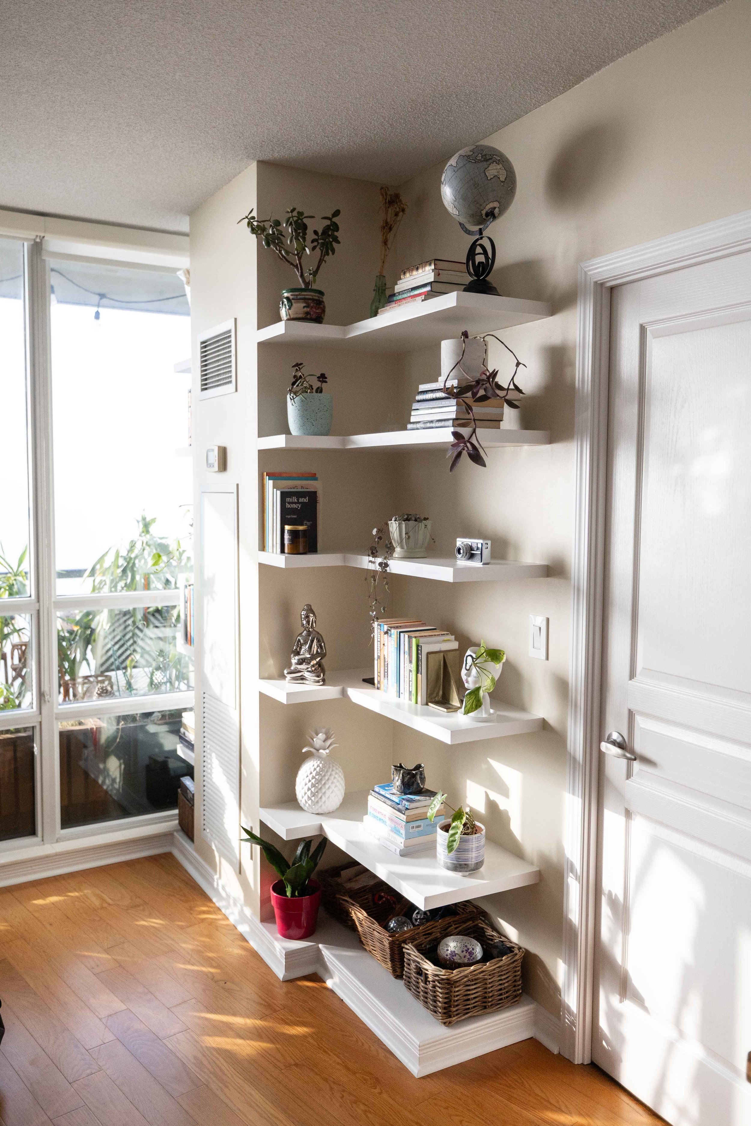 A white wall with a white shelving unit containing books, plants, and decorative items. There is a wicker basket with eggs on the bottom shelf, and a tall potted plant in a red pot on the floor. A sliding door leads to a balcony or outdoor space with