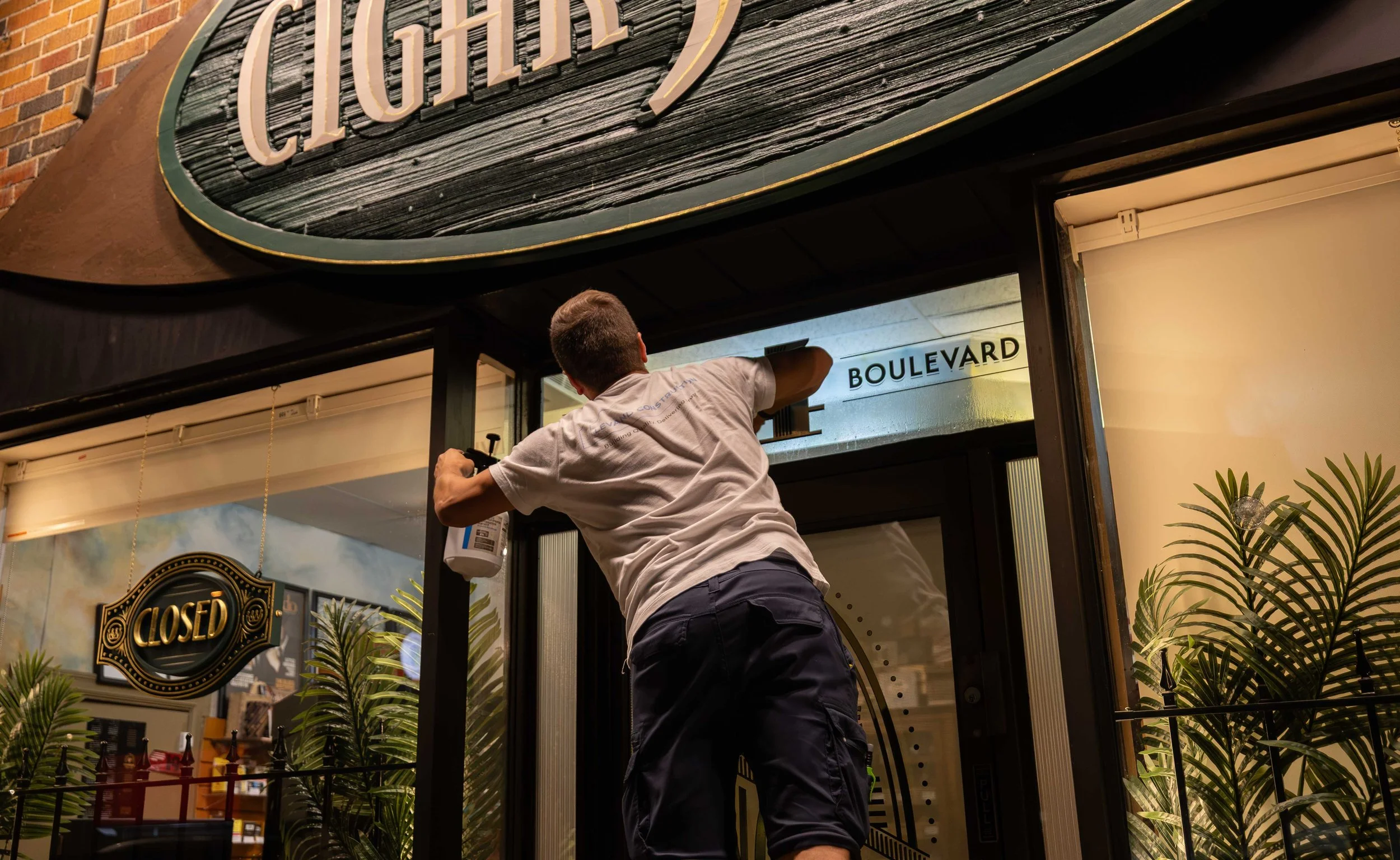 A person with short hair in a gray t-shirt adjusting a door sign labeled "BOULEVARD" at a restaurant entrance.