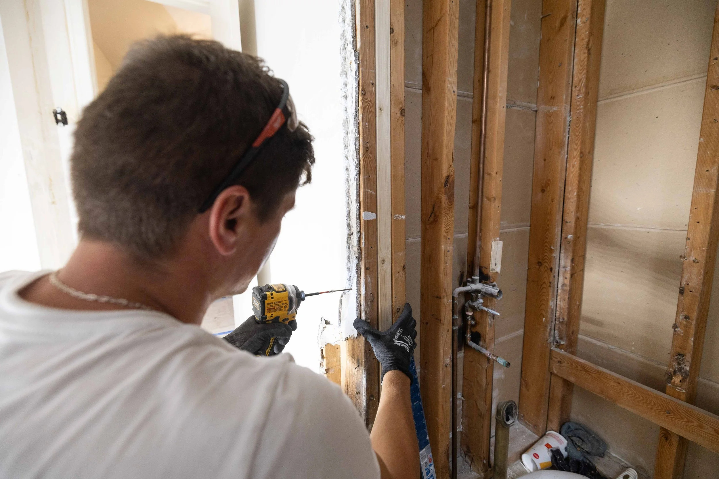 A man wearing a white shirt and black gloves using a drill on wooden framing in a room under construction.