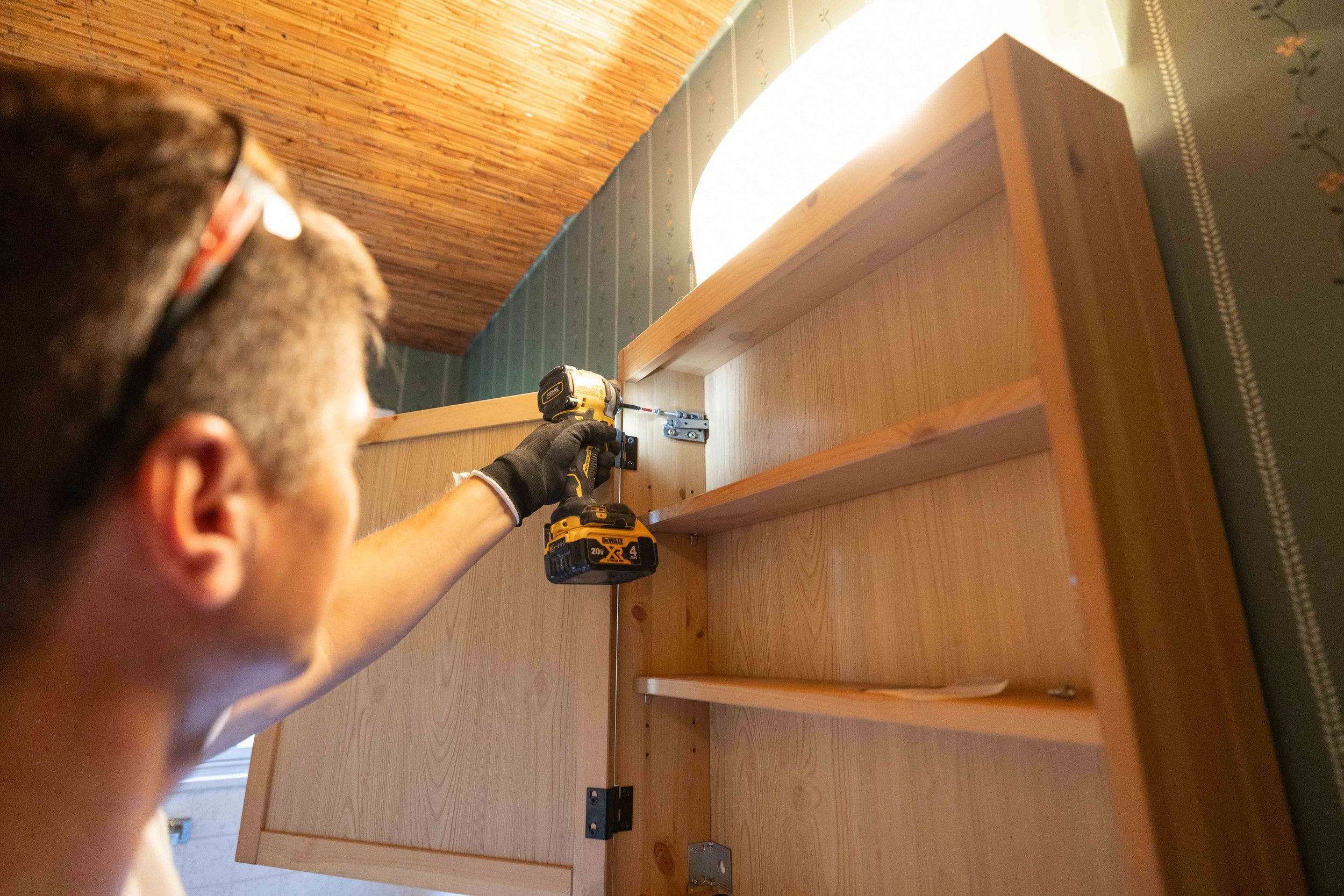 A person using a cordless drill to assemble a wooden piece of furniture in a room with wood-paneled ceiling and decorative wallpaper.