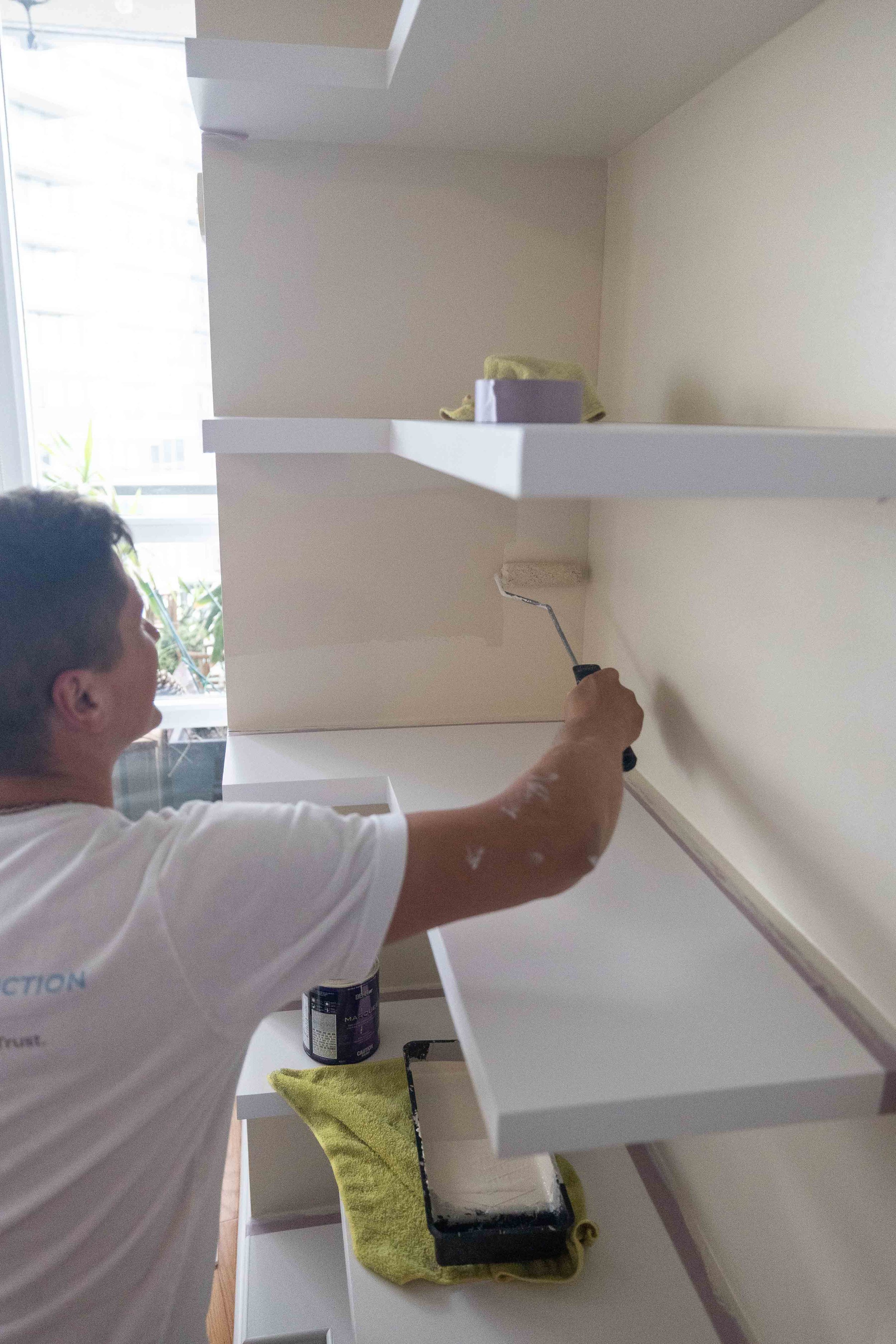A man painting a white shelf with a paint roller in a bright room, using a container of dark paint and a yellow towel nearby.