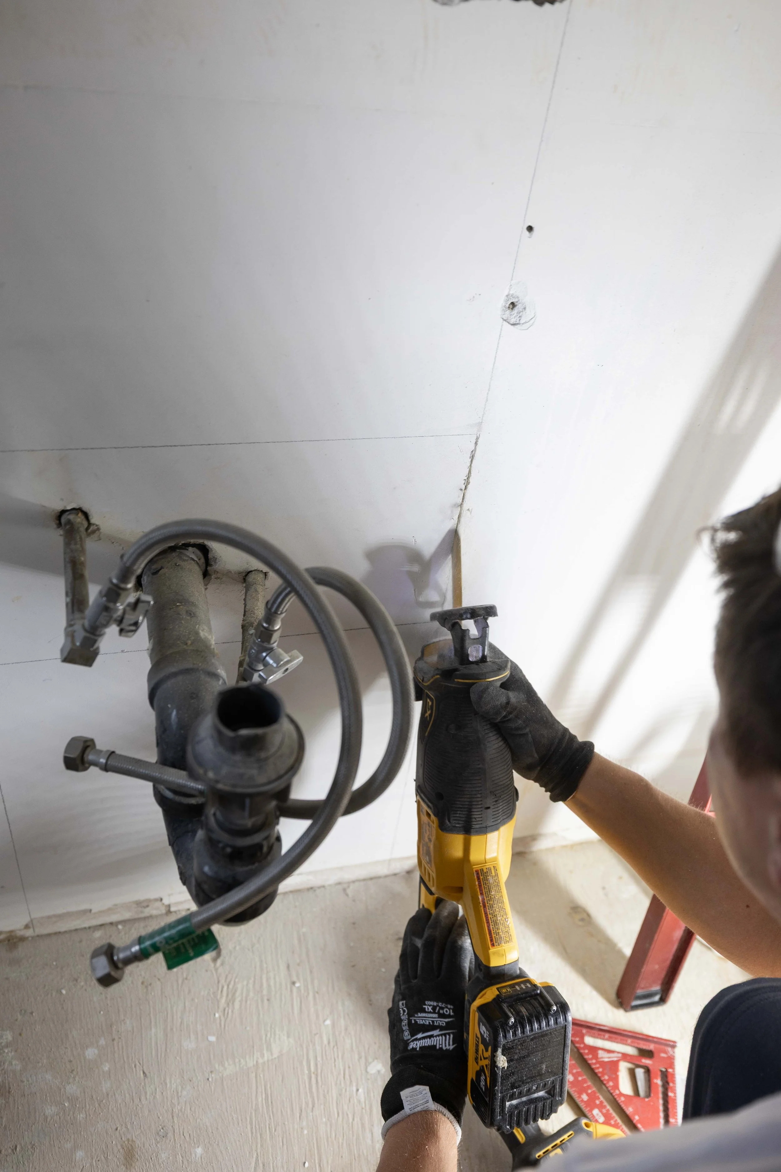 Person using a cordless drill to work on plumbing pipes under a kitchen sink, with a ladder and tools nearby.