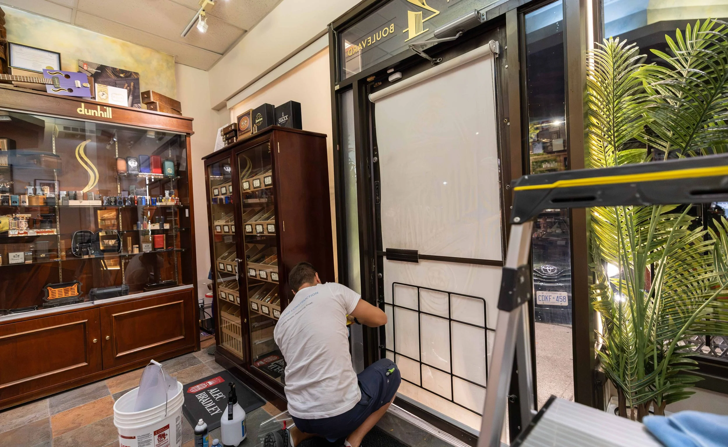 A man is working on assembling a large display board or frame inside a storefront, with shelves of merchandise, guitars, and a plant nearby.