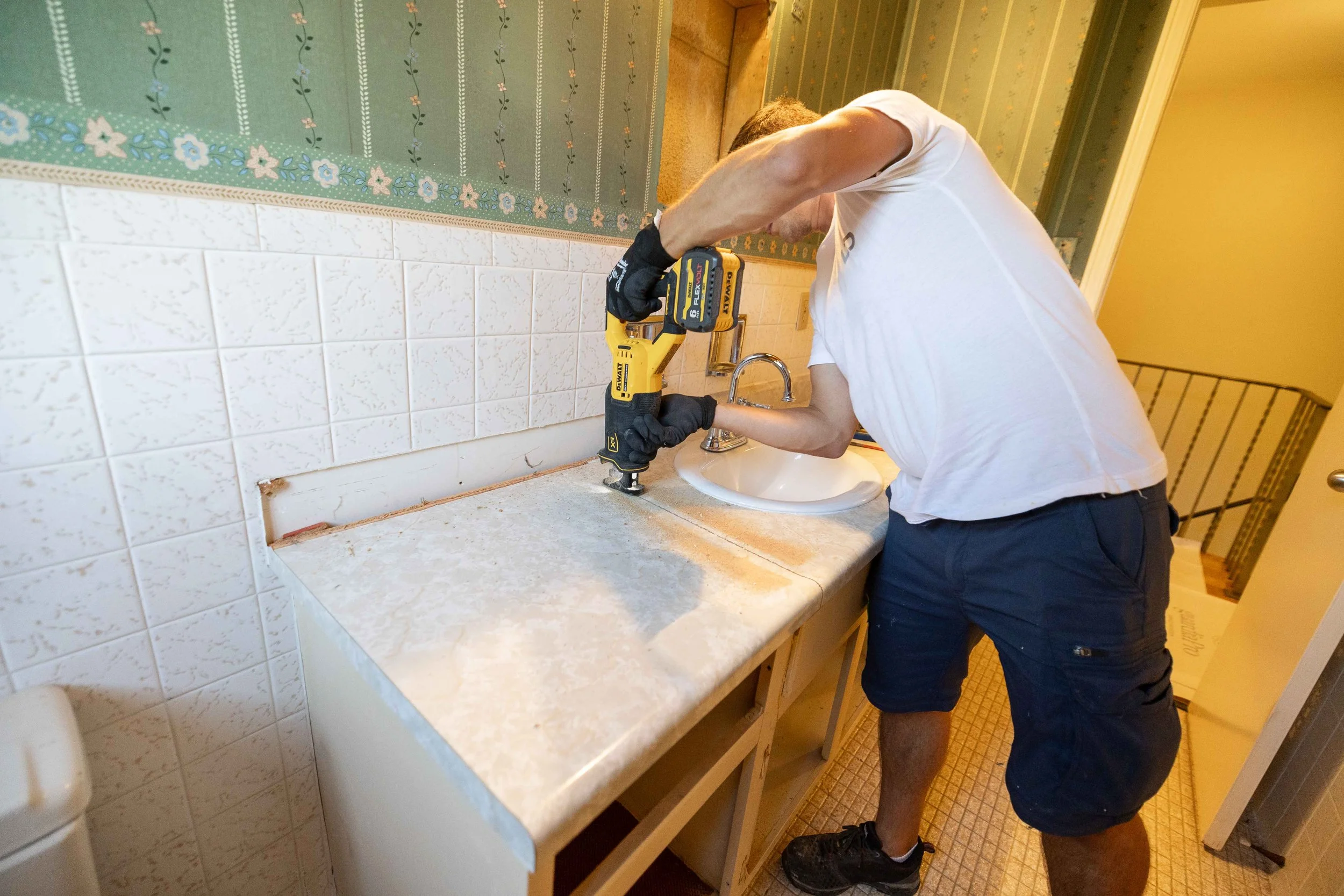 A man wearing a white t-shirt, black shorts, gloves, and a hair band is working on a kitchen countertop with a yellow power drill. The kitchen has white tiled walls, a sink, and green wallpaper with a floral pattern.