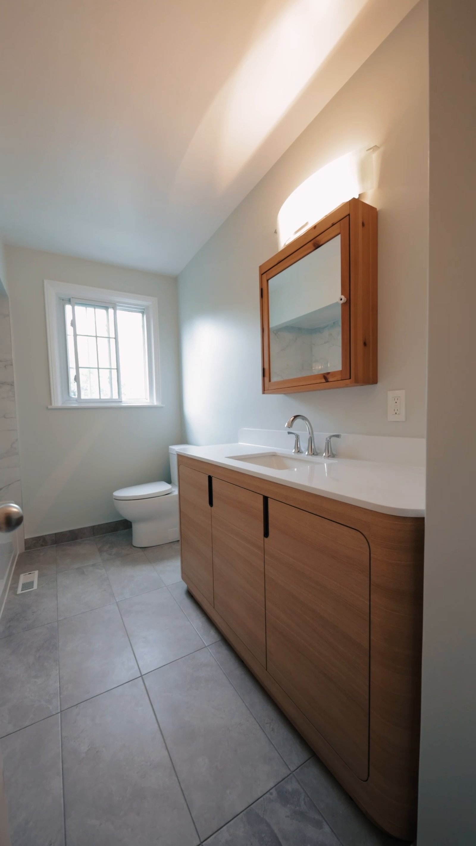 Finished bathroom renovation in Etobicoke with a new wood vanity, white countertop, large grey floor tile and a bright window