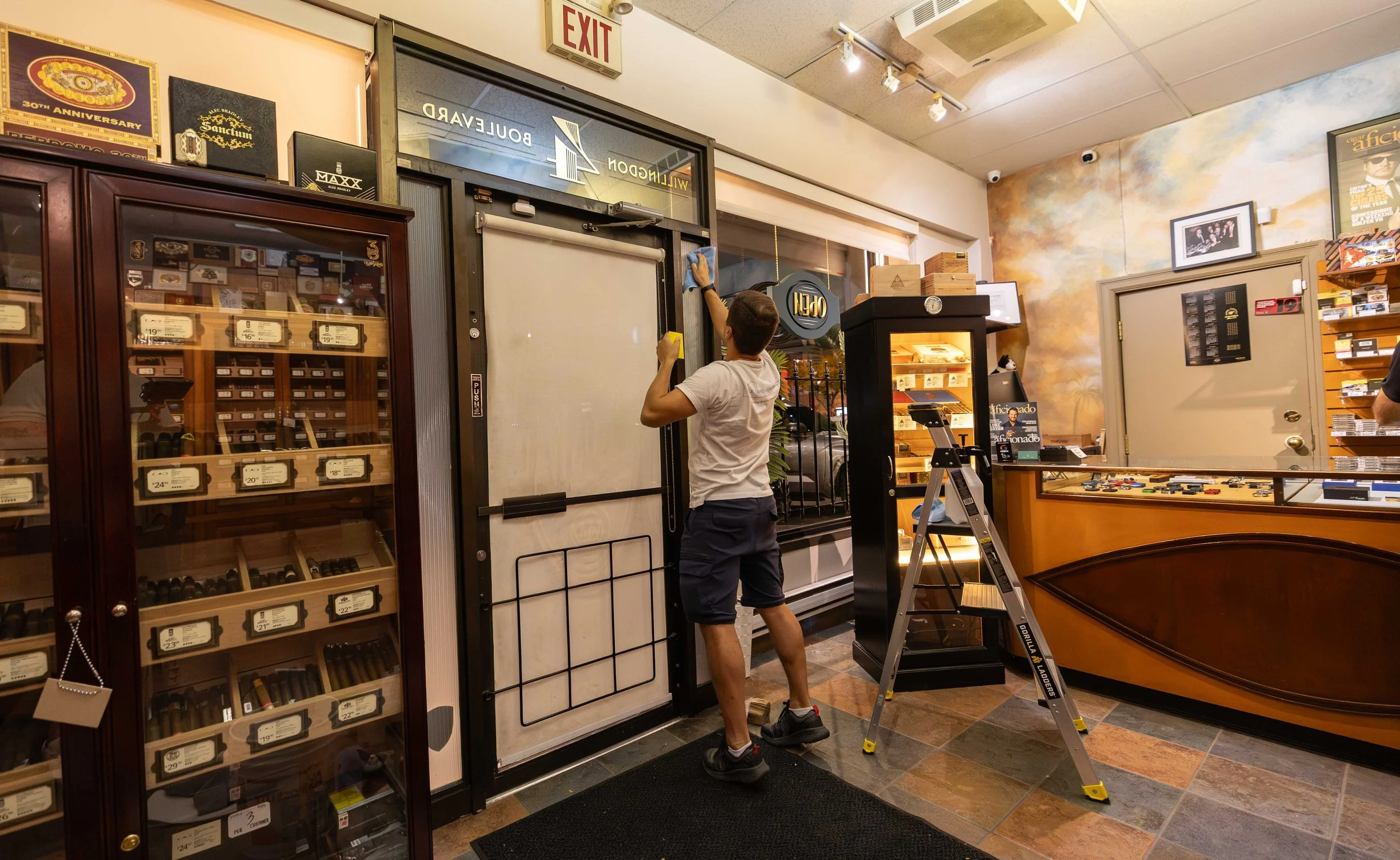 A person cleaning a door in a store, with shelves of cigars and posters on the walls.