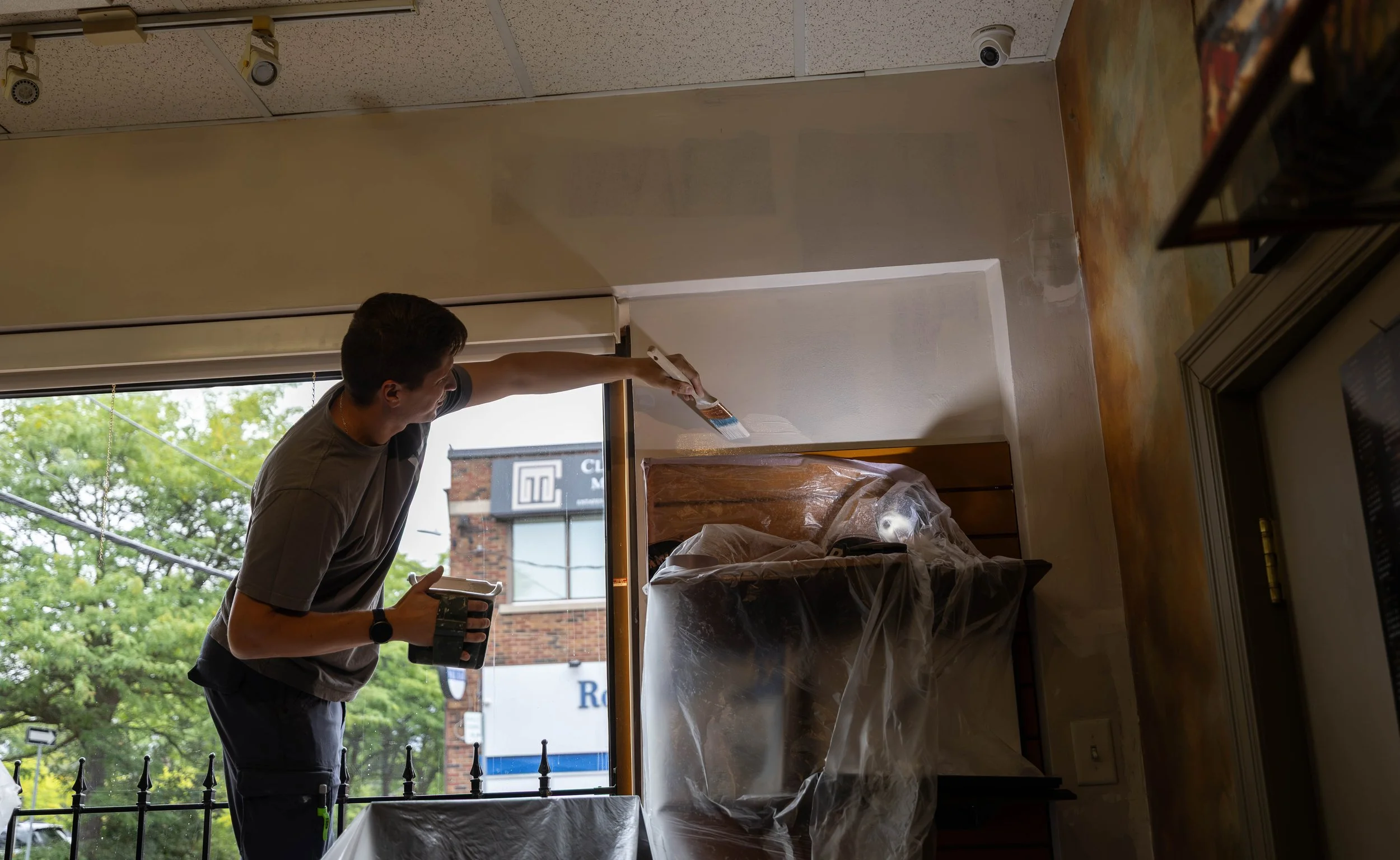 A man painting a wall with a paintbrush in a room with large windows and a view of trees outside.