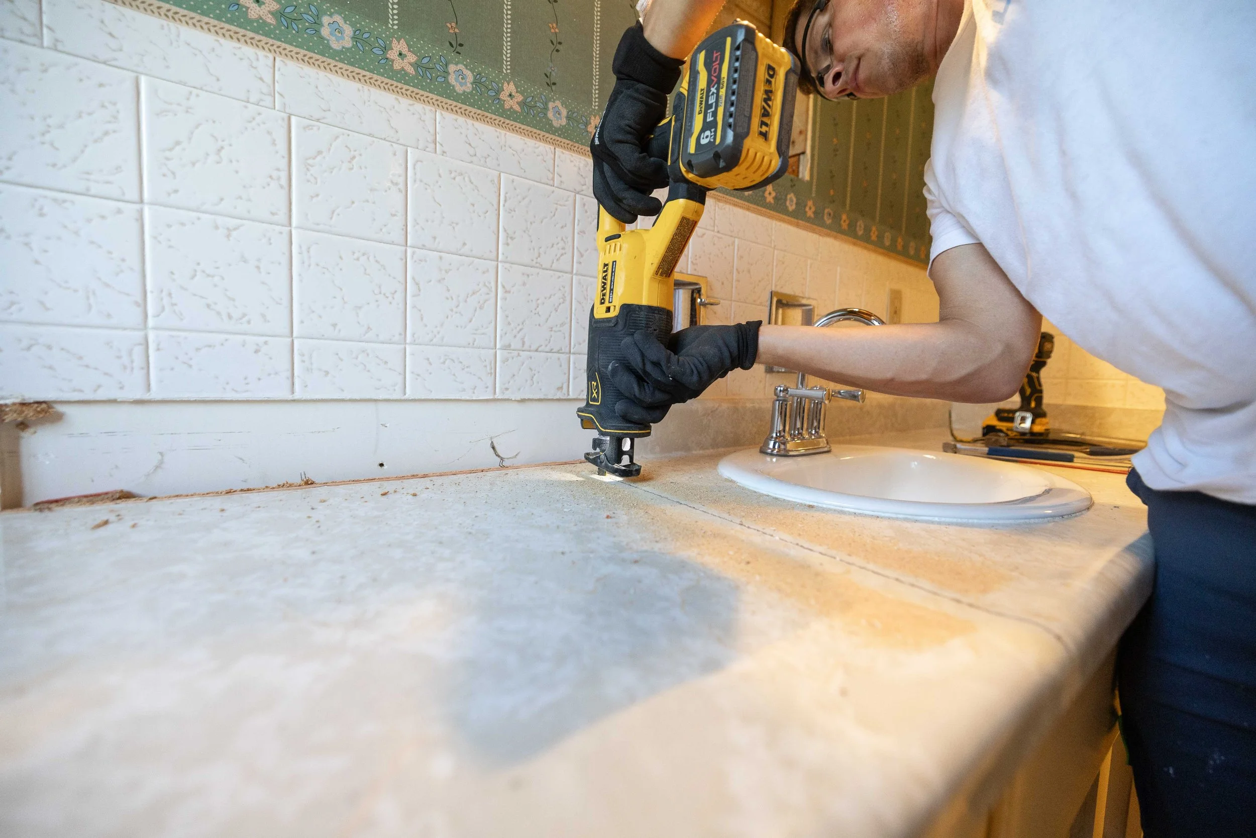 A man in a white t-shirt and glasses uses a yellow DeWalt cordless drill to install or repair a subway tile backsplash above a kitchen sink, with tools and a decorative wallpaper visible in the background.