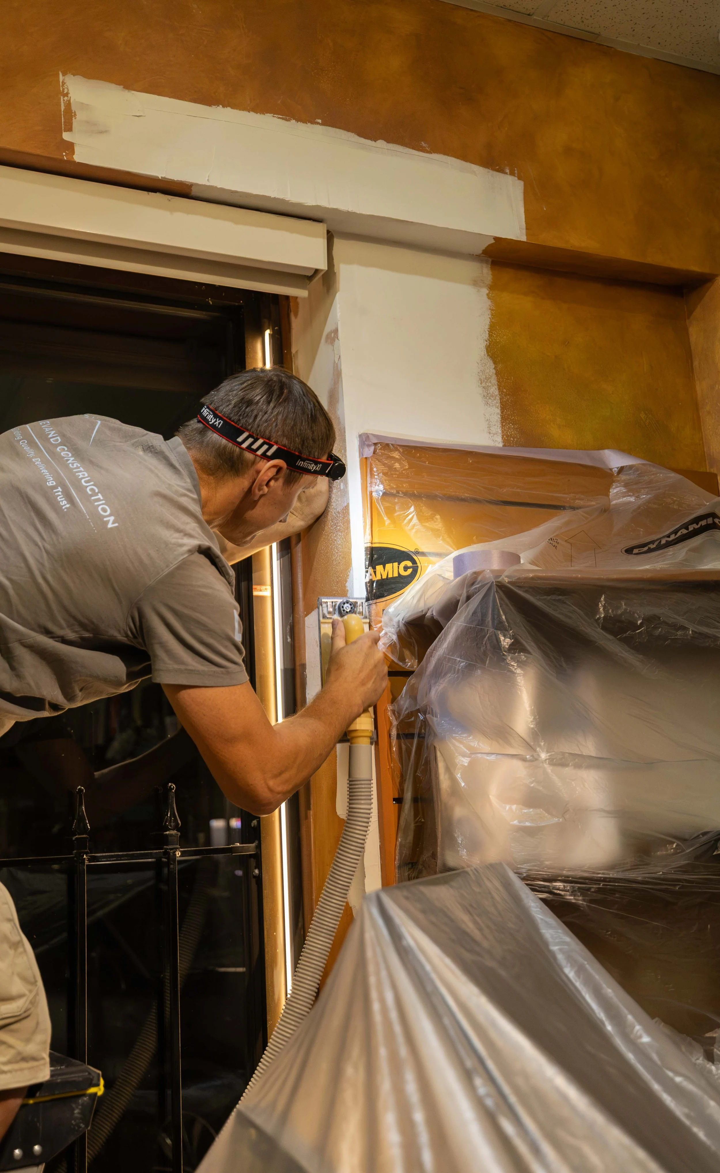 A person working on painting or repair near a window, with plastic sheeting covering furniture or objects, and wearing safety glasses.