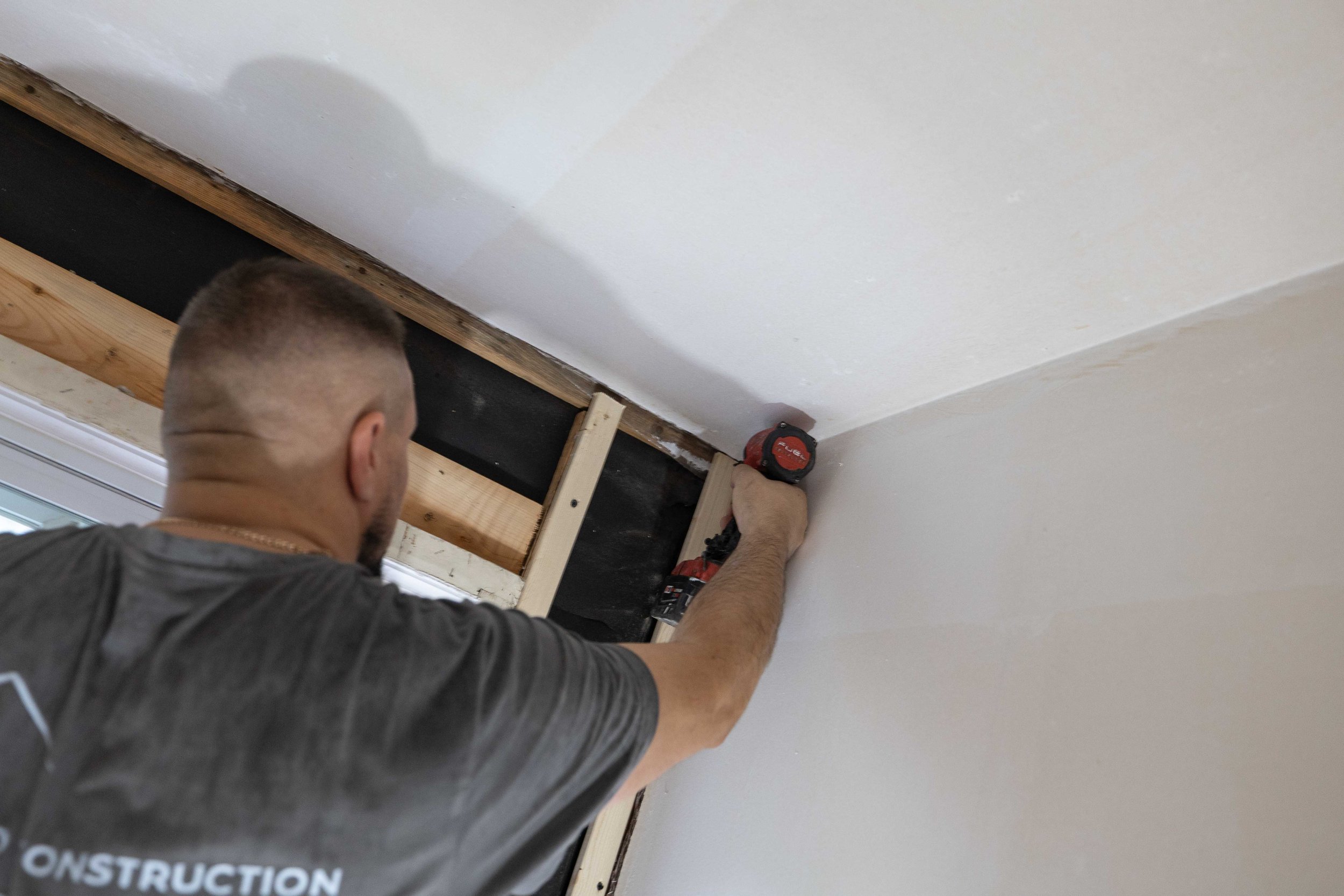 A man installing or repairing a wooden frame or support near the ceiling of a room with drywall, using a power drill.