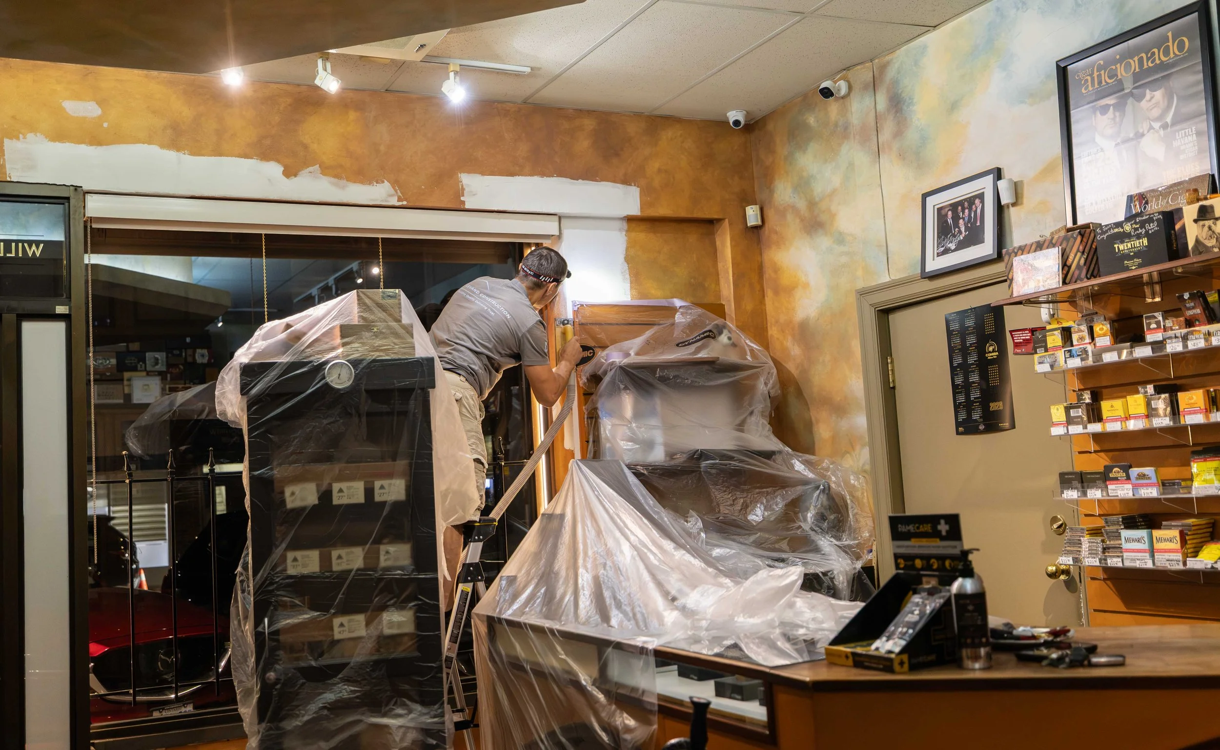 A person working on painting a wall inside a store, surrounded by furniture and shelves, with plastic covering items and equipment.