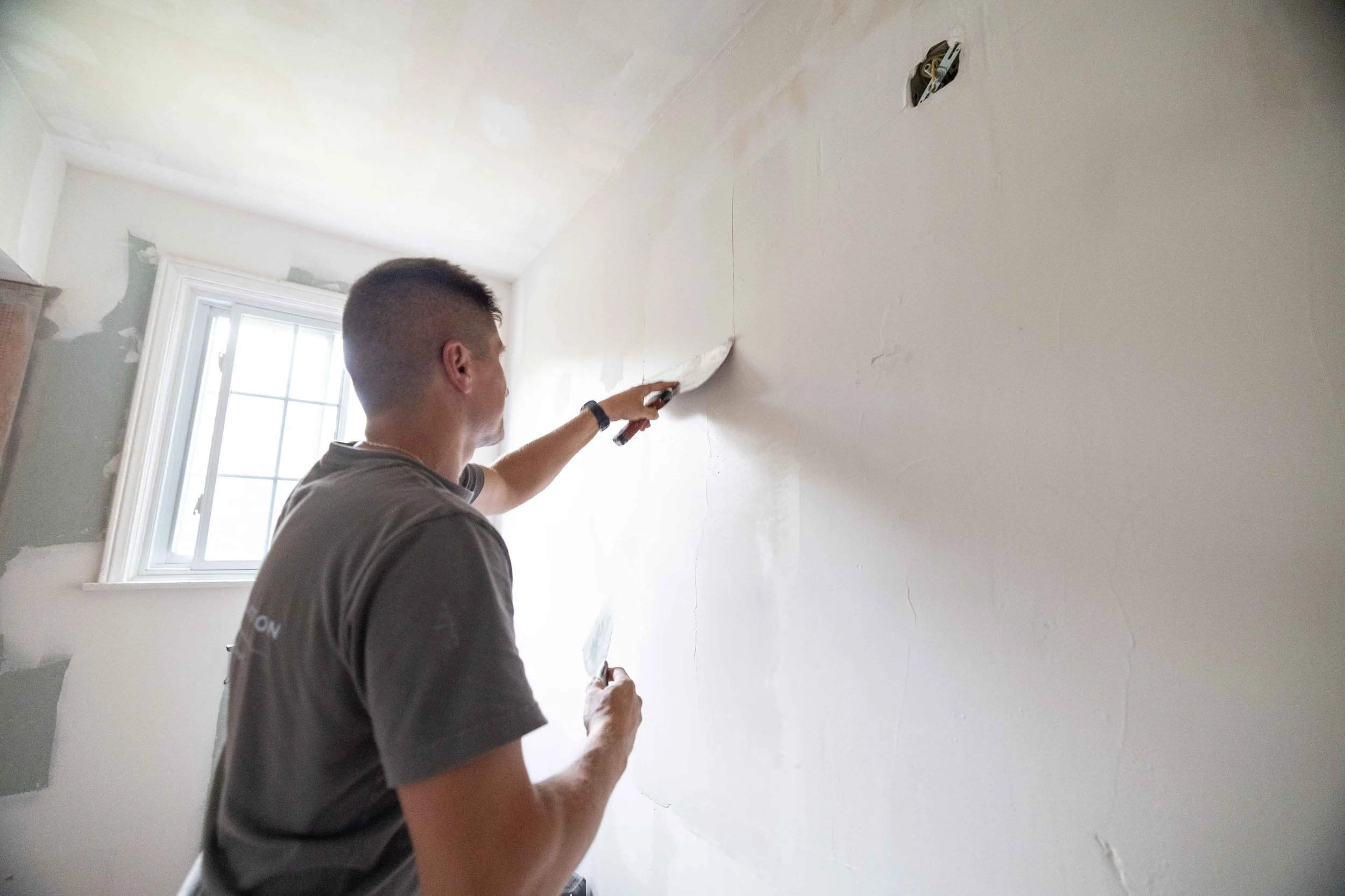 A man working on drywall in a room with a window, using a putty knife to apply putty or joint compound to the wall.