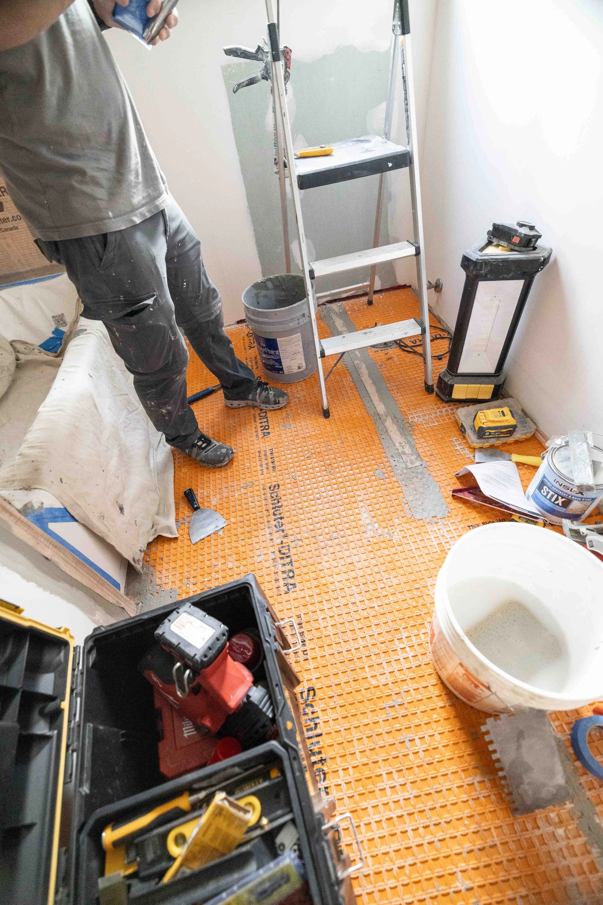 A room under renovation with construction tools, a step ladder, and flooring materials visible. A man in work clothes stands on the left holding a phone. The floor is covered with orange safety padding, and various tools and supplies are scattered ar